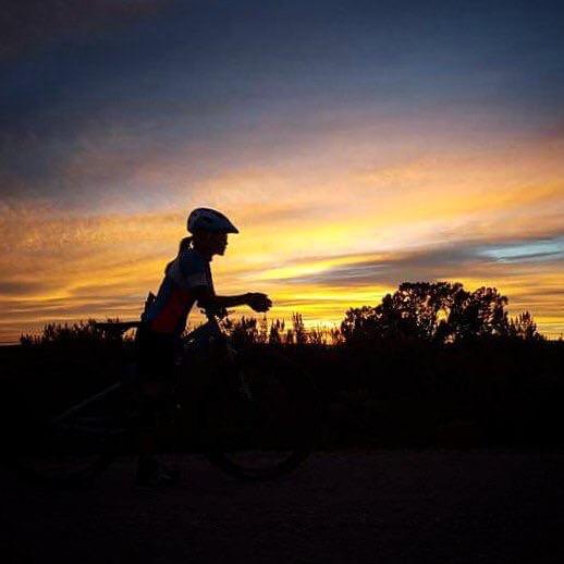 A silhouette of a cyclist riding a mountain bike at dusk, with a vibrant sunset in the background displaying shades of orange, pink, and purple, and dark outlines of trees on the horizon. 18 Road Trails / North Fruita Desert mountain bike trail.