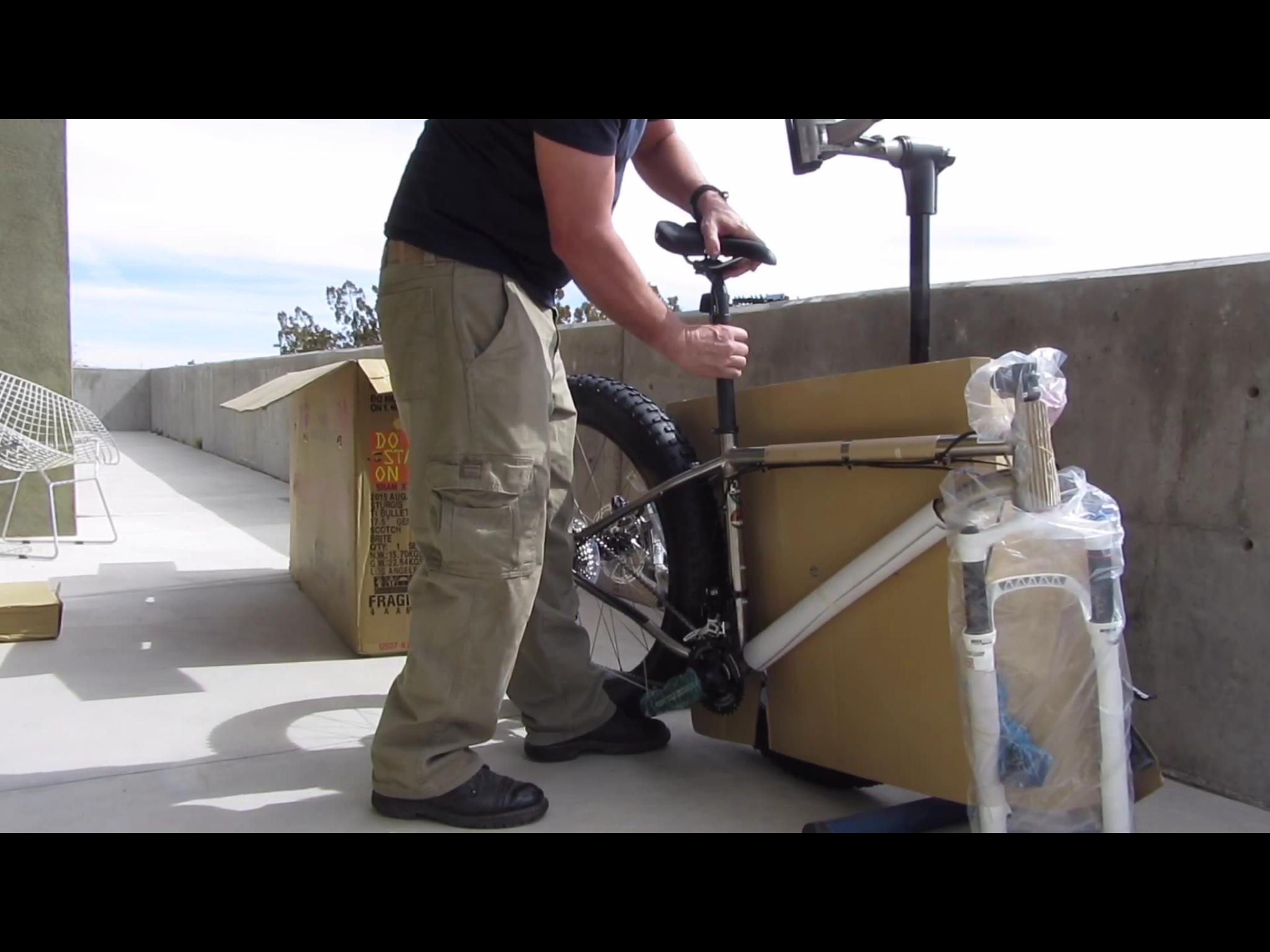 Motobecane Motobecane Bullet Titanium Fat bike: A person assembling a bicycle on an outdoor patio, using tools to adjust the bike's seat. A partially opened cardboard box is nearby, containing bike parts. The scene includes a concrete floor and a few scattered items, with a clear sky in the background.
