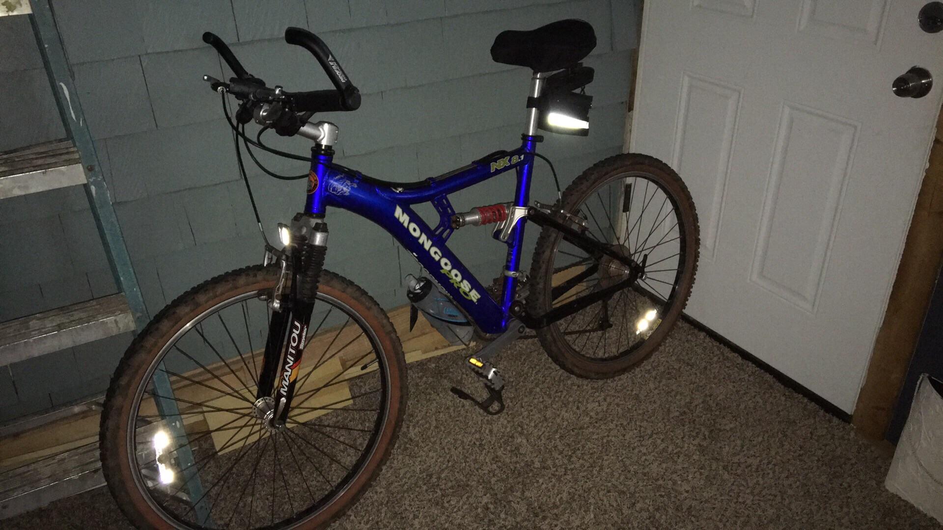 Mongoose MONGOOSE XR-PRO: A blue Mongoose mountain bike parked next to a white door in a dimly lit room. The bike features front suspension and reflectors on the tires. A metal ladder and wooden planks are visible in the background. The floor is covered in carpet.