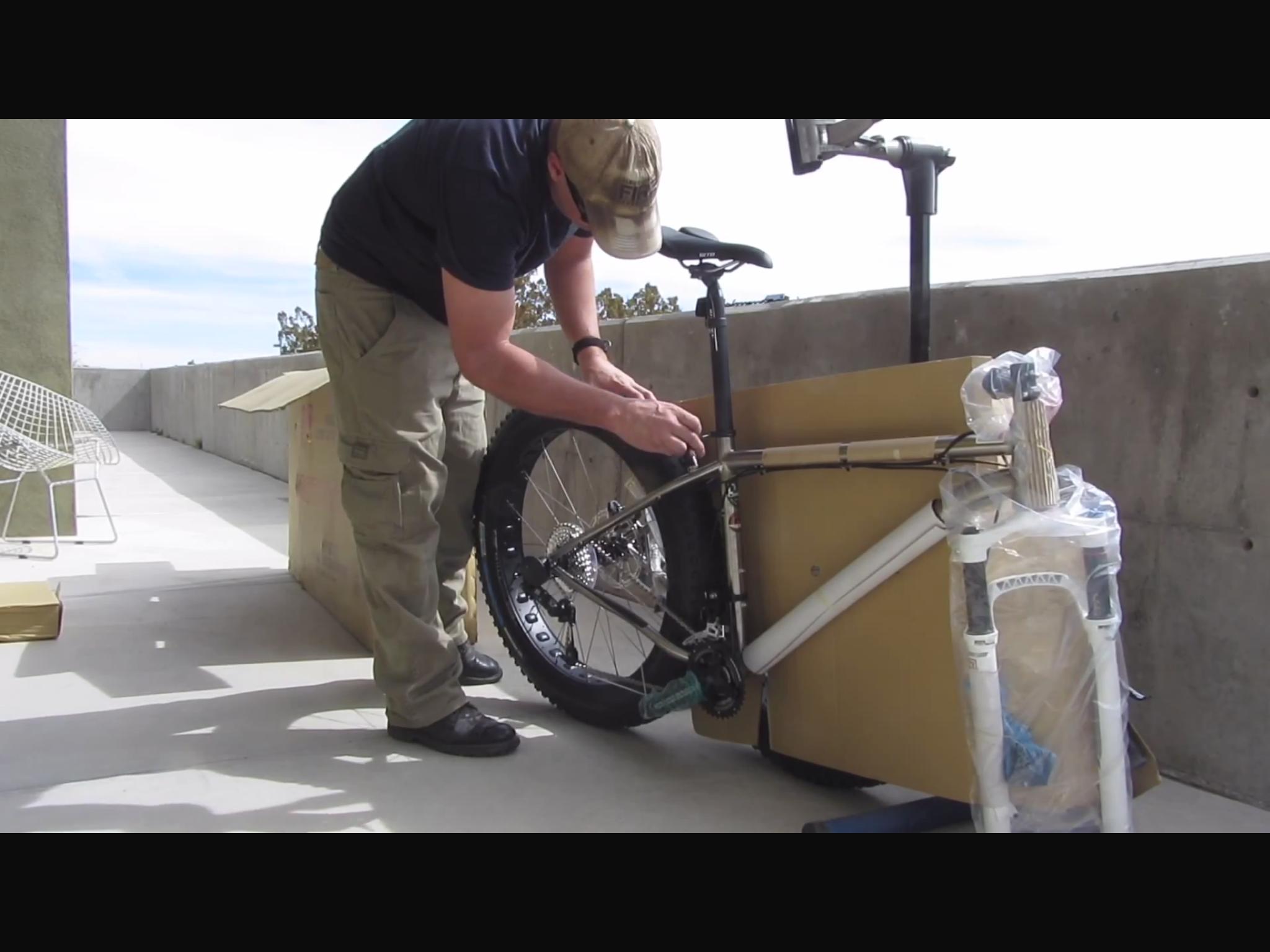 Motobecane Motobecane Bullet Titanium Fat bike: A person is assembling a bicycle on a balcony, with parts partially unpacked from boxes. The individual is adjusting the rear wheel of the bike, which has a large tire and is secured against a cardboard box. The scene is set outdoors, with a clear sky visible in the background.