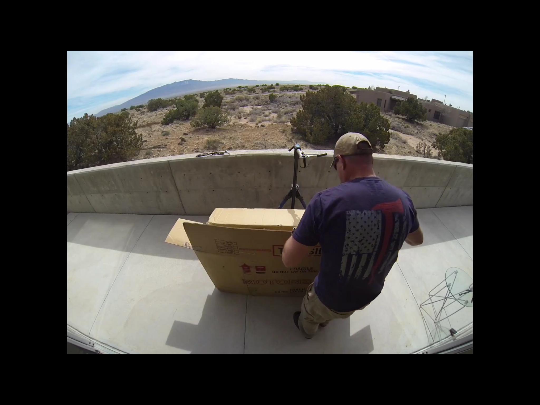 Motobecane Motobecane Bullet Titanium Fat bike: A person wearing a hat and a T-shirt with an American flag design is unpacking a large cardboard box on a concrete balcony. The outdoor setting features a desert landscape with shrubs and distant mountains under a partly cloudy sky. A tripod and a wire structure are visible nearby.