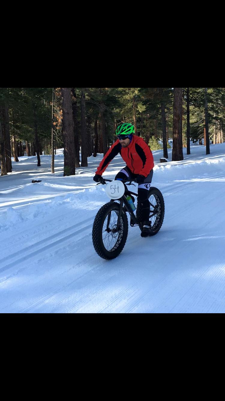 A person riding a fat bike on a snowy trail in a forest. The cyclist is wearing a green helmet and a red jacket, with their focus directed forward. Snow-covered trees line the path, and the ground is blanketed in snow, showcasing a winter landscape. The bike has wide tires designed for snow, and a number plate labeled "54" is attached to the front. Burton Creek State Park mountain bike trail.