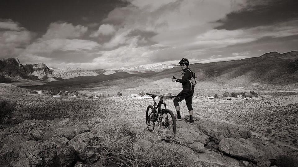 A mountain biker stands on a rocky outcrop, overlooking a vast, arid landscape with distant mountains and a small settlement. The scene is captured in black and white, highlighting the textures of the rocks and clouds in the sky. The biker, dressed in cycling gear and a helmet, holds a device while standing next to their mountain bike. Cottonwood Valley North mountain bike trail.