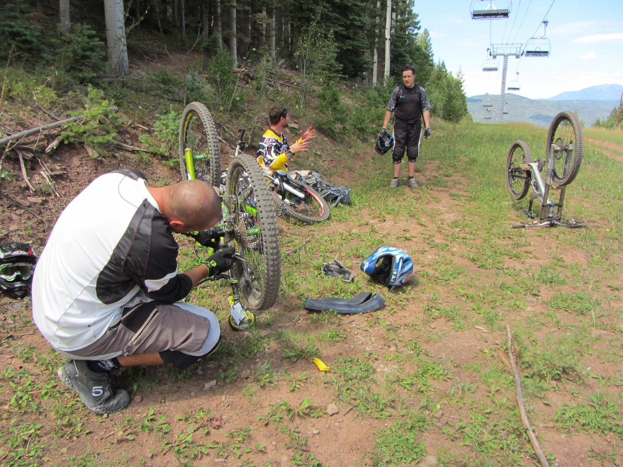 Three mountain bikers in a forested area are working on their bikes. One rider, kneeling and wearing a white and black jersey, is repairing a bicycle wheel, while another rider stands nearby in black gear, looking on. The third bike is lying upside down on the ground. Several helmets and biking gear can be seen scattered around them, and a ski lift is visible in the background. Angel Fire Bike Park mountain bike trail.