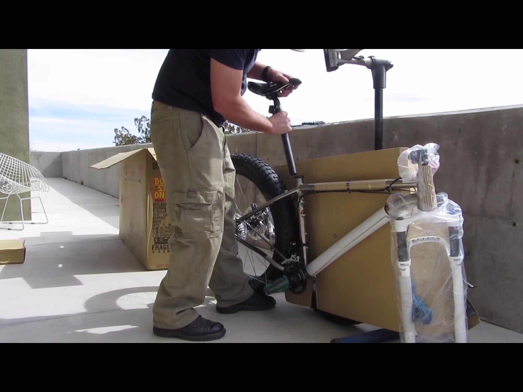 Motobecane Motobecane Bullet Titanium Fat bike: A person wearing a black t-shirt and cargo pants is assembling a mountain bike on a balcony. The bike is partially unpacked, with its frame visible and protective plastic still on some components. A cardboard box is nearby, and the background features a concrete wall and a chair. The sky is clear with some clouds.