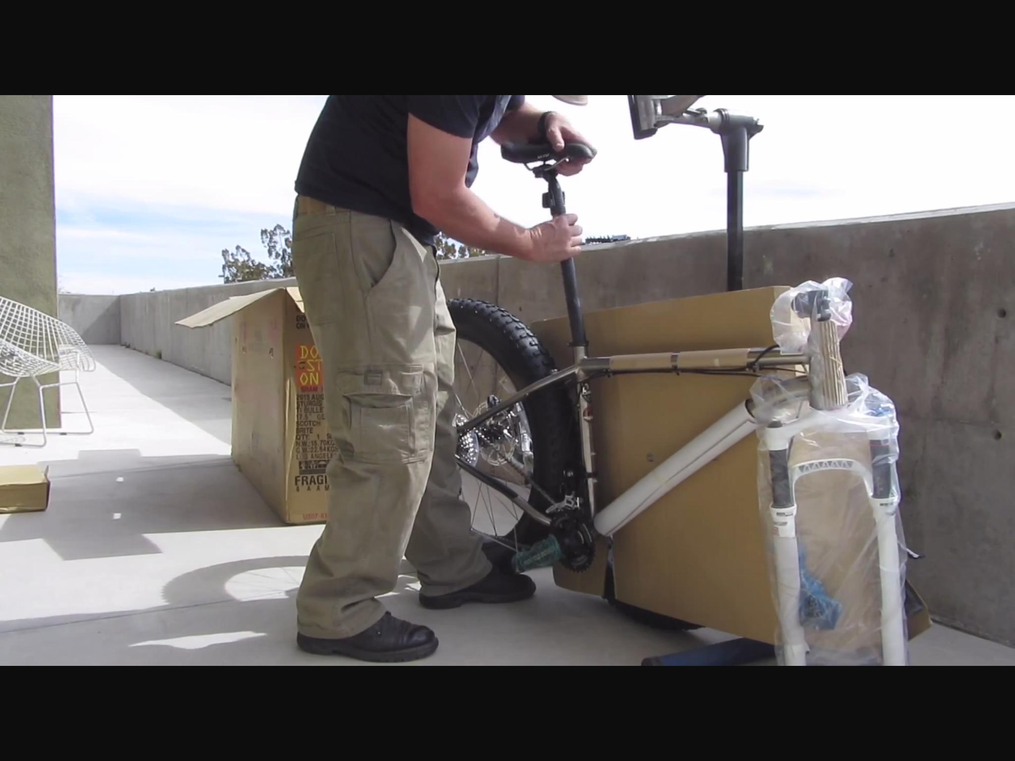 Motobecane Motobecane Bullet Titanium Fat bike: A person assembling a mountain bike on a balcony, using a bike stand. The bike is partially unpacked, with its frame visible, and some components still wrapped in plastic. A cardboard box sits nearby, indicating that the bike was just delivered. The background features a concrete wall and a piece of outdoor furniture.