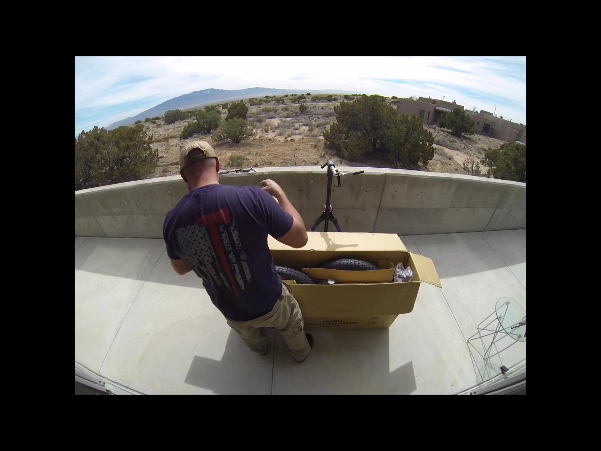 Motobecane Motobecane Bullet Titanium Fat bike: A person standing on a balcony with their back facing the camera, unpacking a large cardboard box. Inside the box are various parts, including a wheel. The background features a desert landscape with shrubs and distant mountains under a partly cloudy sky.