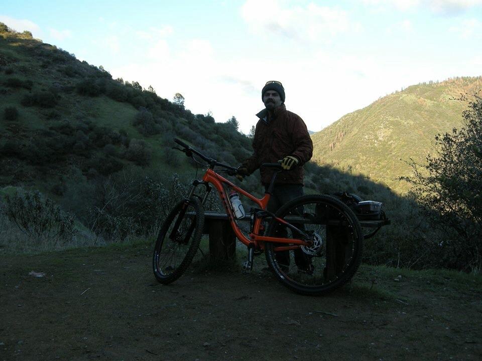 Trek Fuel EX 5 29: A person standing next to an orange mountain bike at a scenic outdoor location, with green hills and trees in the background. The individual is wearing a brown jacket and gloves, and the lighting suggests either early morning or late afternoon.