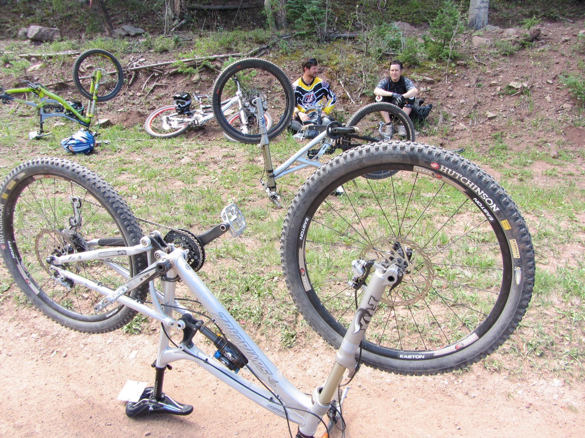 Two mountain bikers resting on the ground beside several overturned mountain bikes. One bike is propped up on its handlebars, while others are scattered in the background. The scene is set in a grassy area surrounded by trees. Angel Fire Bike Park mountain bike trail.