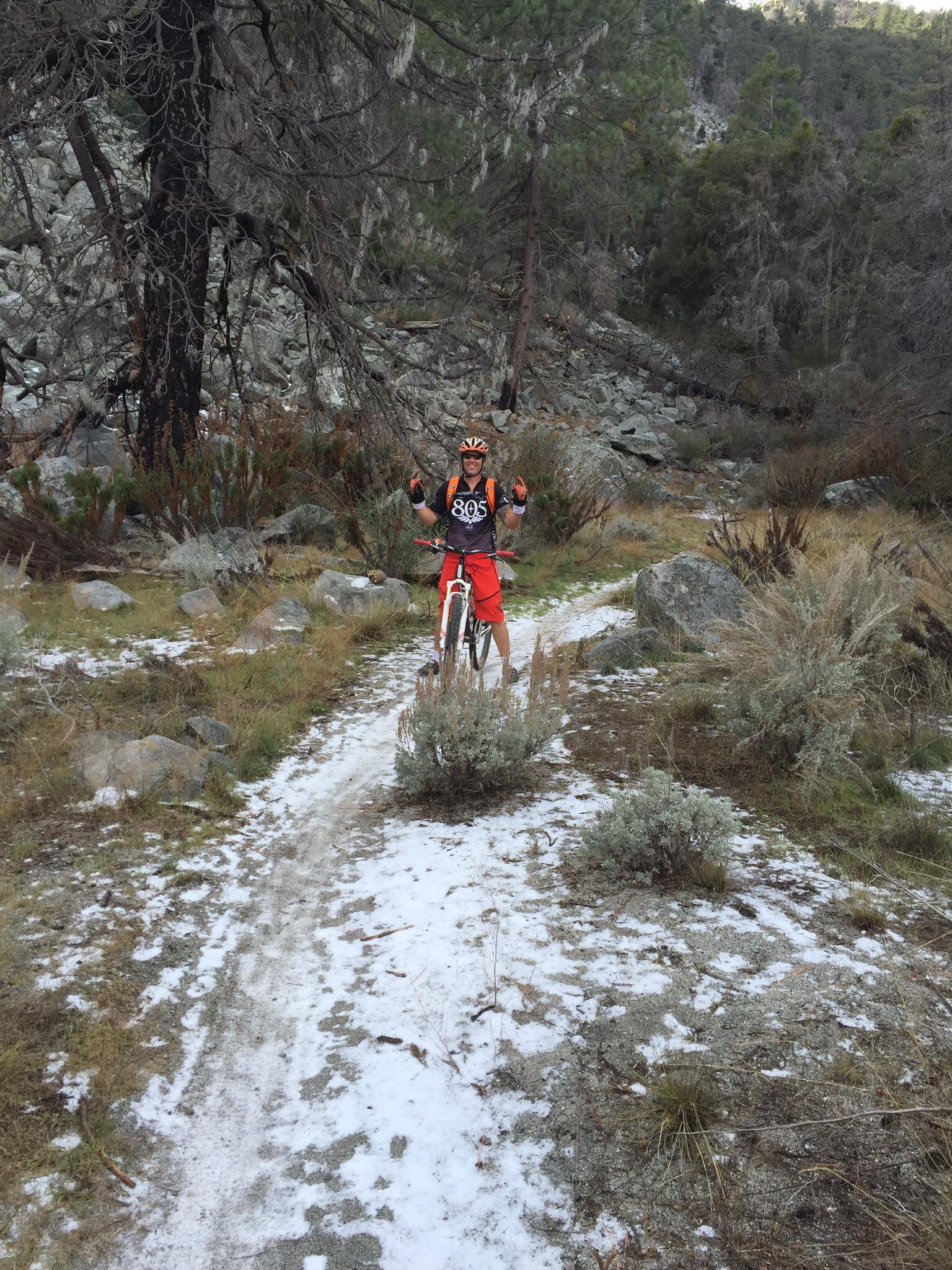 A mountain biker in a black and orange outfit poses with a thumbs-up on a rocky trail, surrounded by vegetation and trees. The path appears to have some snow, indicating winter conditions. El Prieto mountain bike trail.