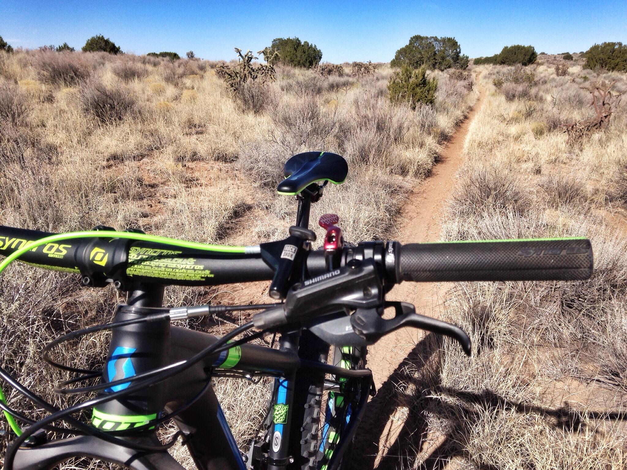 A close-up view of a mountain bike handlebar, showcasing the grips and brake lever, with a dirt trail and sparse vegetation in the background under a clear blue sky. Super Fat Bike Loop mountain bike trail.