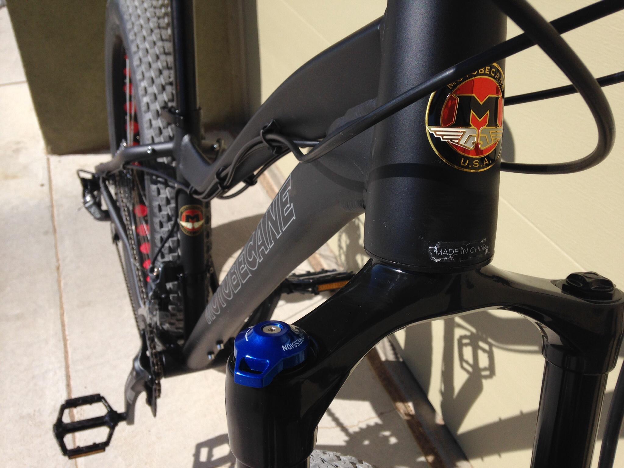 Motobecane Boris: A close-up view of a Motobecane mountain bike showing the frame, fork, and pedals. The bike features a black finish with a visible logo on the headtube and a blue adjustment dial on the fork. Sunlight casts shadows on the ground, highlighting the bike's details.