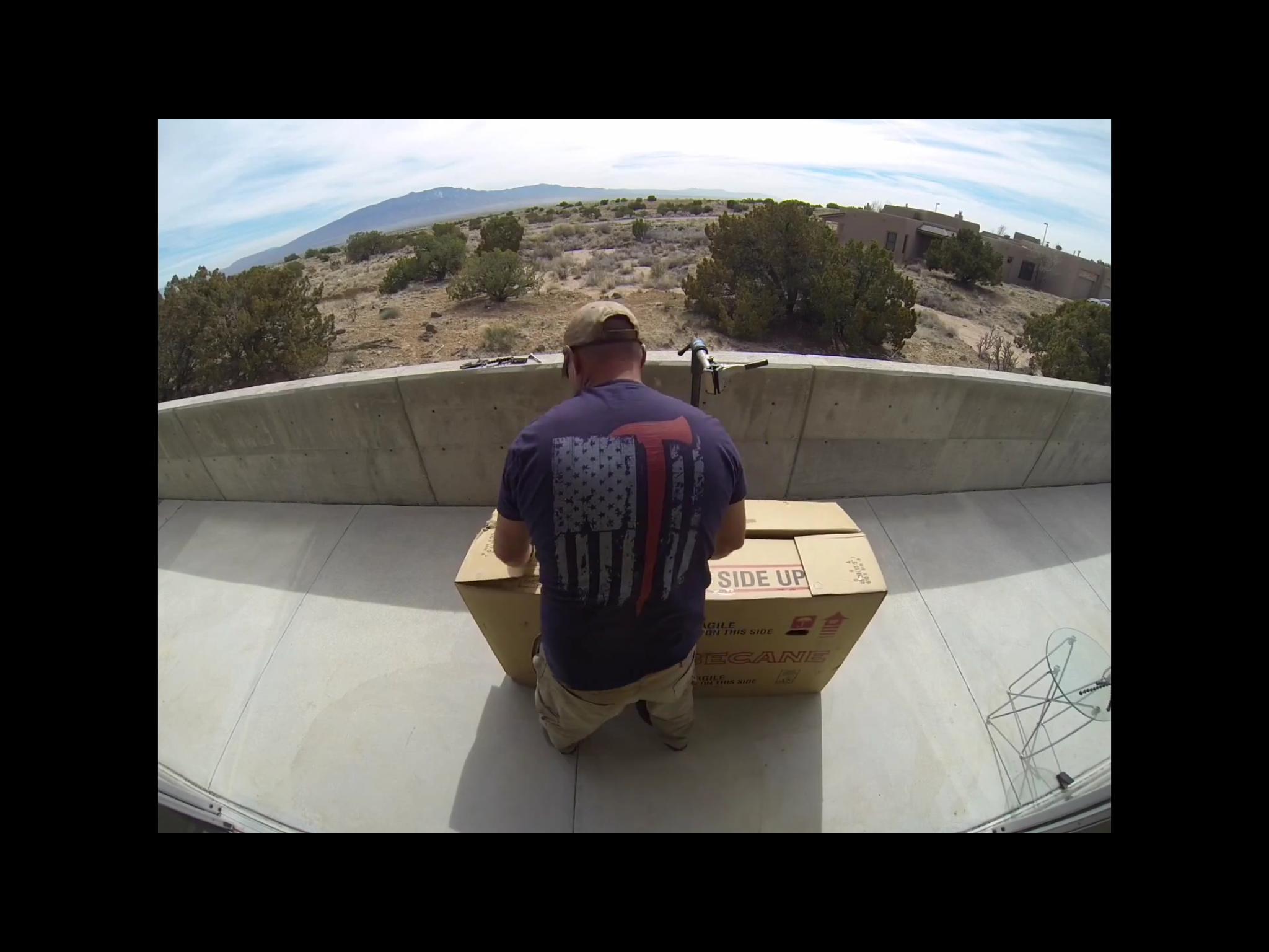 Motobecane Motobecane Bullet Titanium Fat bike: A person wearing a blue shirt with an American flag design is sitting on a patio, preparing to open a large cardboard box labeled "SIDE UP." In the background, there is a desert landscape with shrubs and mountains under a partly cloudy sky.
