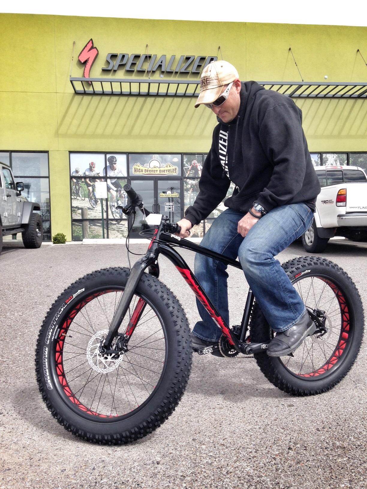 A person dressed in casual attire, wearing a baseball cap and sunglasses, is sitting on a fat bike with large, knobby tires. The background features a green exterior of a bicycle shop with the word "Specialized" prominently displayed. In addition, there are bicycles showcased in the store's windows and a pickup truck parked nearby on a paved surface.