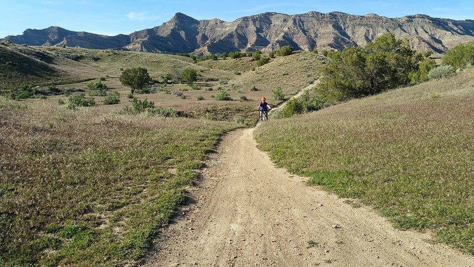A person riding a bicycle on a dirt path through a grassy landscape, with rolling hills and mountains in the background under a clear blue sky. Kessel Run mountain bike trail.