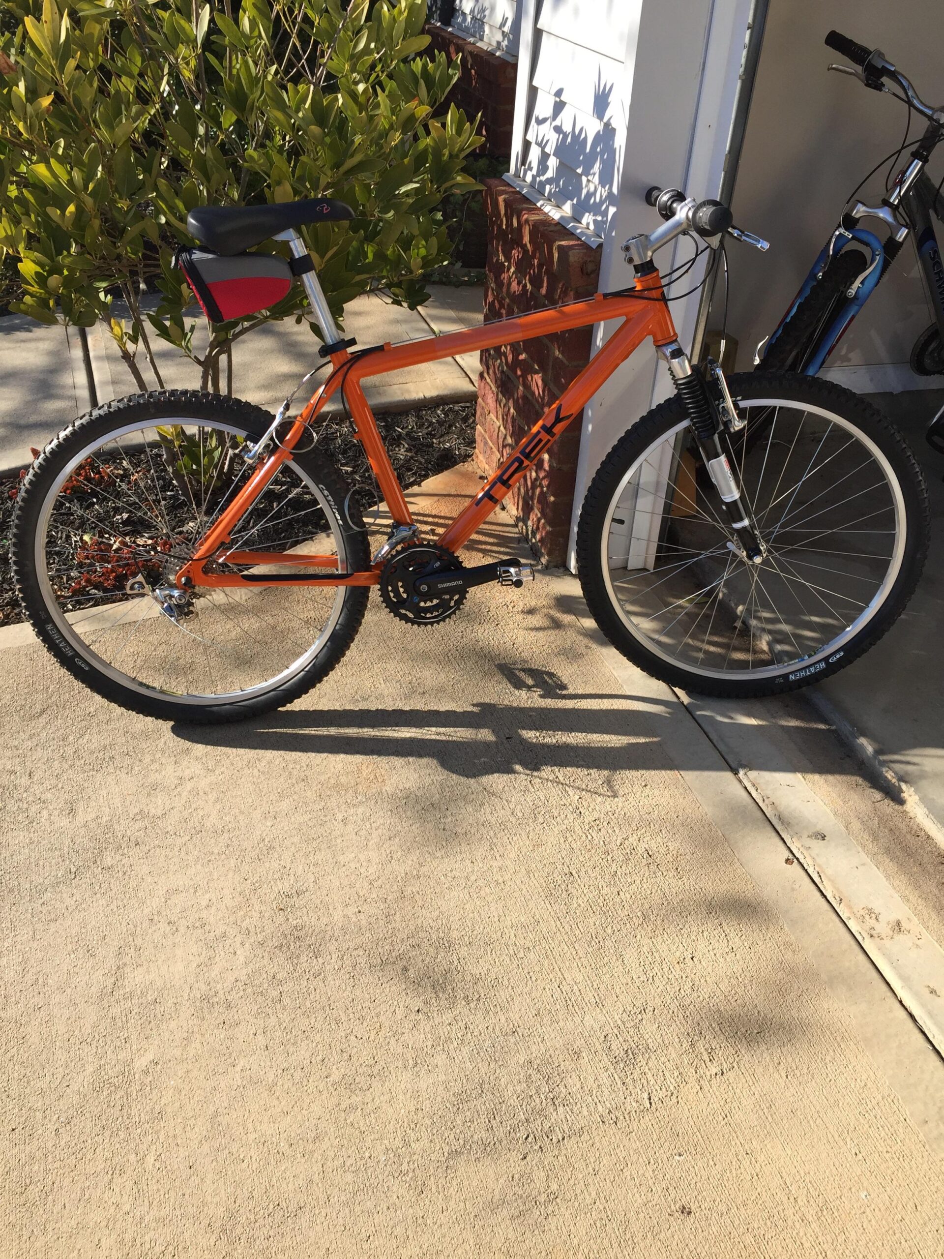 Trek 6000: Alt text: An orange Trek mountain bike with a black saddle and a small red bag attached to the seat, parked on a concrete surface next to a house with green shrubs in the background. Another bike can be seen in the background.