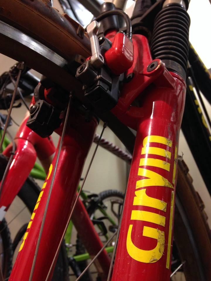Close-up view of the front fork of a red mountain bike featuring the brand name "Girvin." The image highlights the brake and suspension components, with a blurred background showing additional bikes.