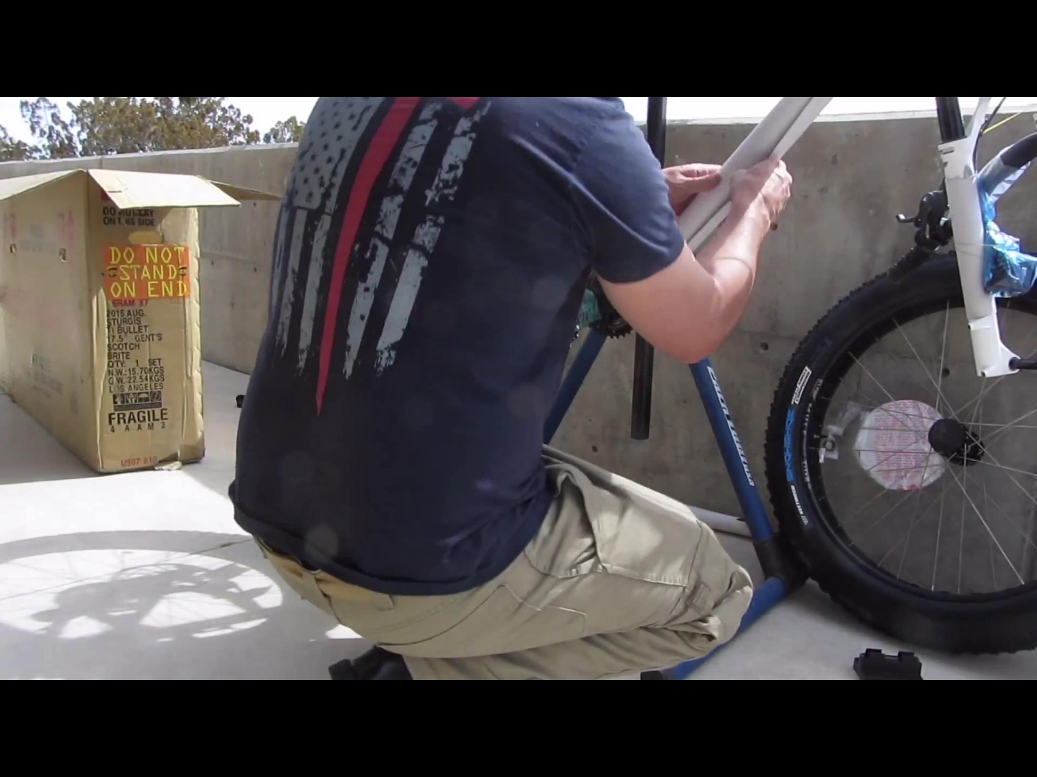 Motobecane Motobecane Bullet Titanium Fat bike: A person kneeling on a surface, working on a bicycle frame, which is being held in a bike stand. A cardboard box with "DO NOT STAND ON END" written on it is in the background. The person is wearing a dark shirt with a graphic design and light-colored pants. The scene suggests a bike assembly or maintenance task outdoors.