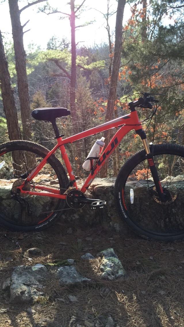 A vibrant orange mountain bike parked on a stone wall in a forested area. The bike features thick tires, a water bottle attached to the frame, and is surrounded by tall trees with green and autumn-colored foliage. Sunlight filters through the trees, illuminating the scene. Forest City Trail mountain bike trail.