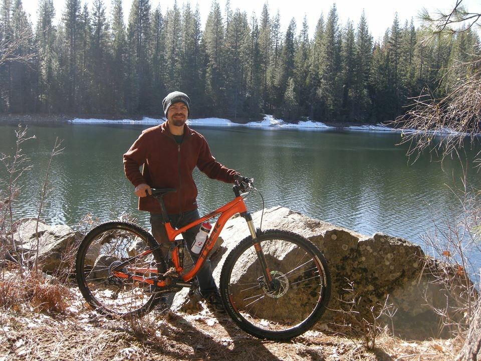 Trek Fuel EX 5 29: A person standing next to an orange mountain bike by a serene lake surrounded by pine trees, with a touch of snow visible in the background. The sun is shining, creating a bright and inviting atmosphere.
