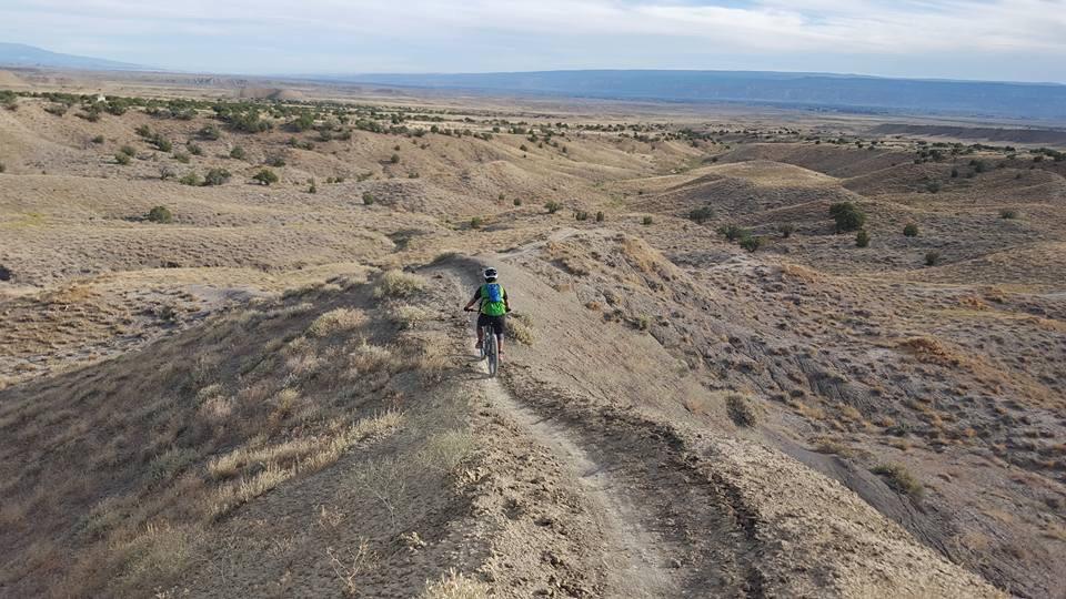 A person riding a mountain bike along a narrow dirt trail that winds through a hilly and arid landscape. The scene features rolling hills with sparse vegetation under a cloudy sky, showcasing a remote outdoor setting ideal for cycling. Joe's Ridge mountain bike trail.