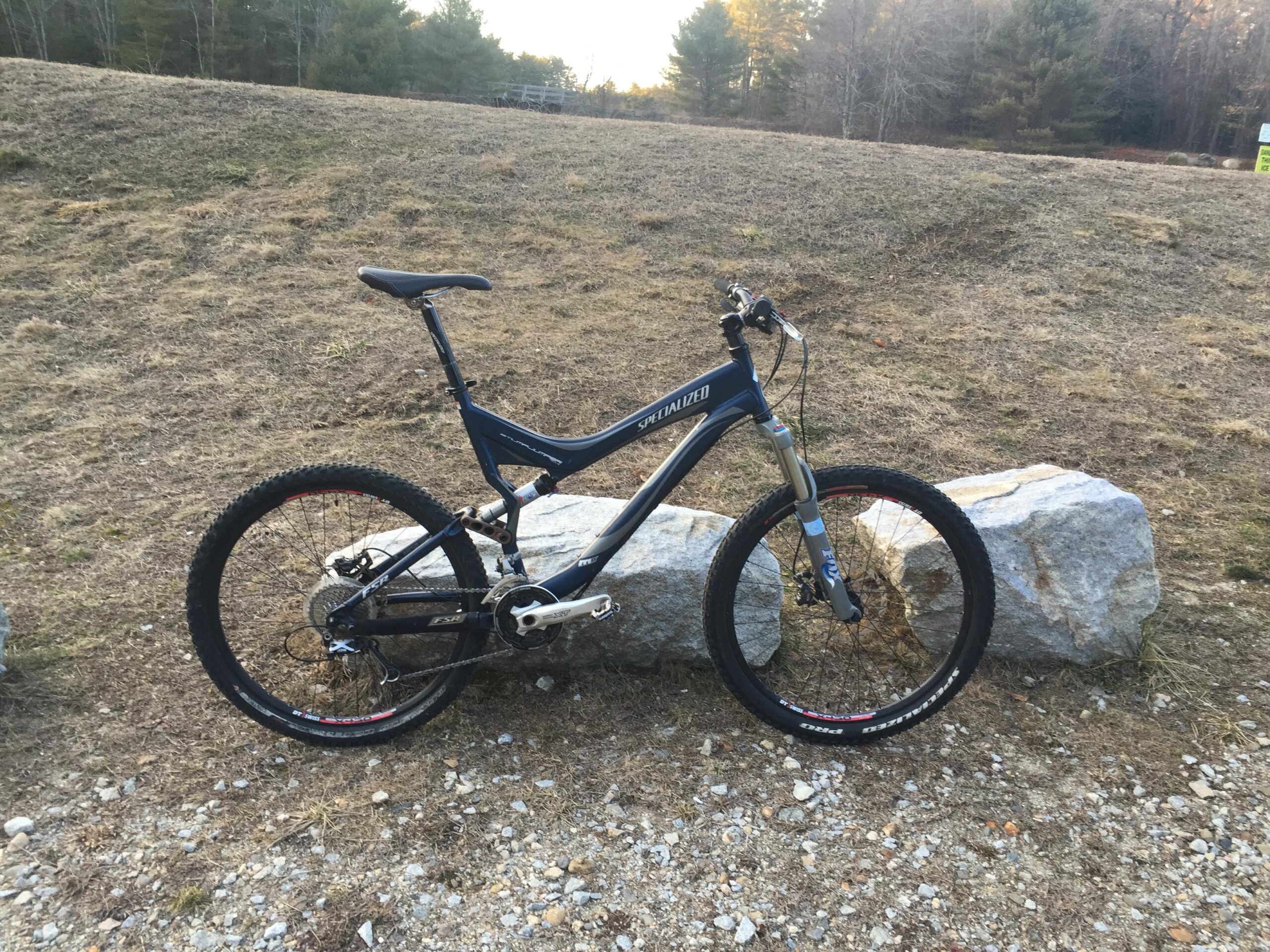Specialized Stumpjumper FSR Elite: A blue mountain bike leaning against a large rock, set against an open grassy area with sparse trees in the background. The bike features thick tires designed for off-road use and a lightweight frame.