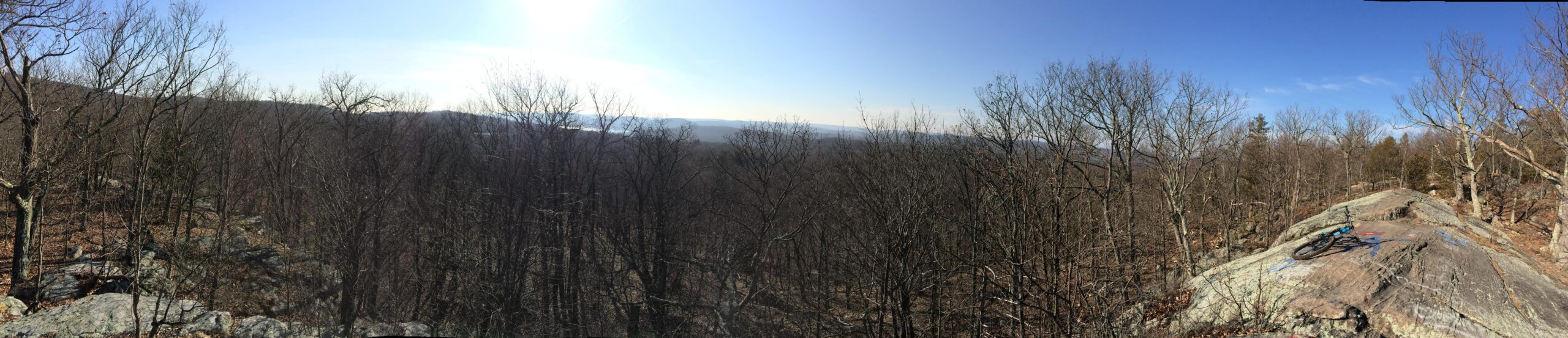 A panoramic view from a rocky outcrop overlooking a forested landscape. The scene features bare trees with a backdrop of distant hills under a clear blue sky. Sunlight illuminates the area, casting shadows on the ground. A mountain bike is positioned on the rock, suggesting an outdoor adventure or biking activity. Ringwood Skylands Manor mountain bike trail.