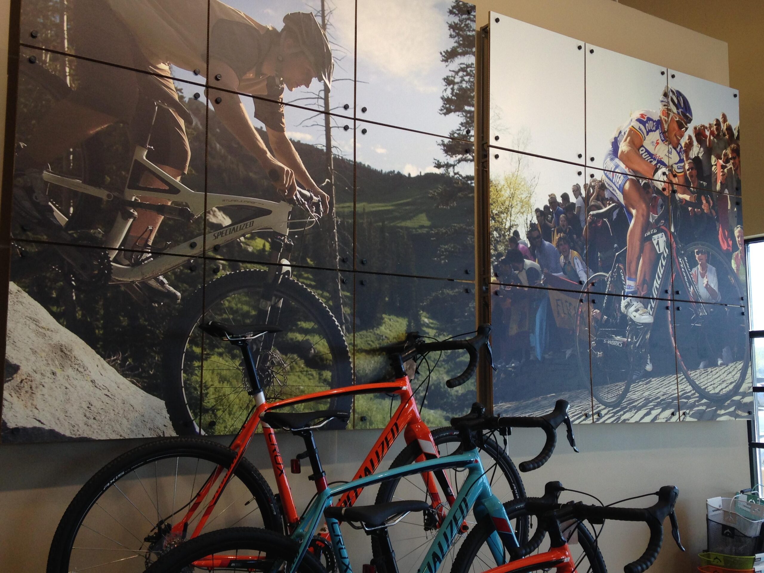 A display featuring two large photographic panels of cyclists in action, one showcasing a mountain biker navigating a rocky terrain and the other depicting a road cyclist surrounded by cheering spectators. In the foreground, two bicycles are visible, one in teal and orange and the other in black, resting against the wall.