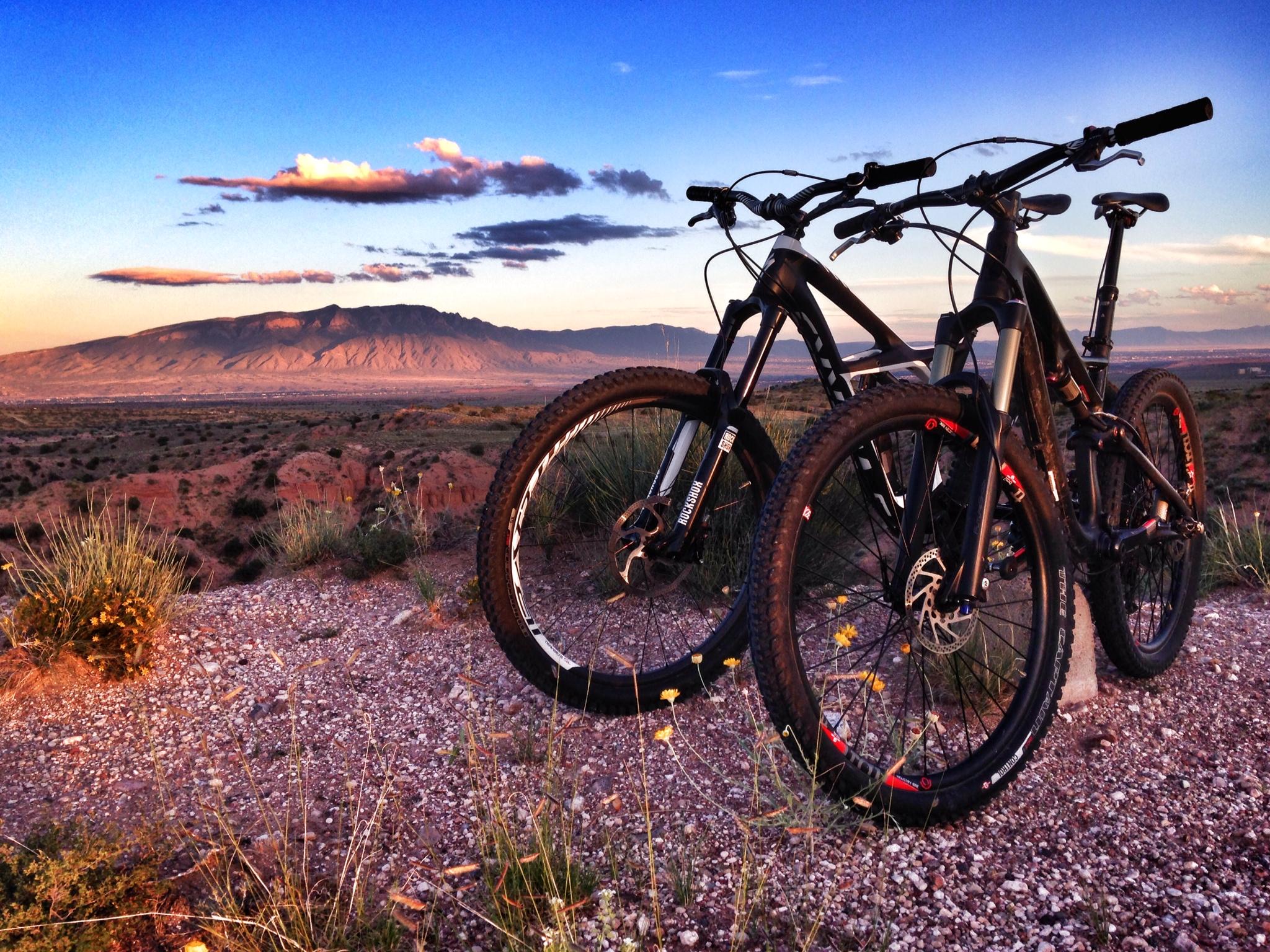Specialized Stumpjumper FSR Expert: Two mountain bikes are parked on a gravel hillside, overlooking a picturesque landscape of rolling hills and distant mountains under a vibrant sunset sky filled with clouds. Wildflowers and grasses surround the bikes, adding a natural touch to the scene.
