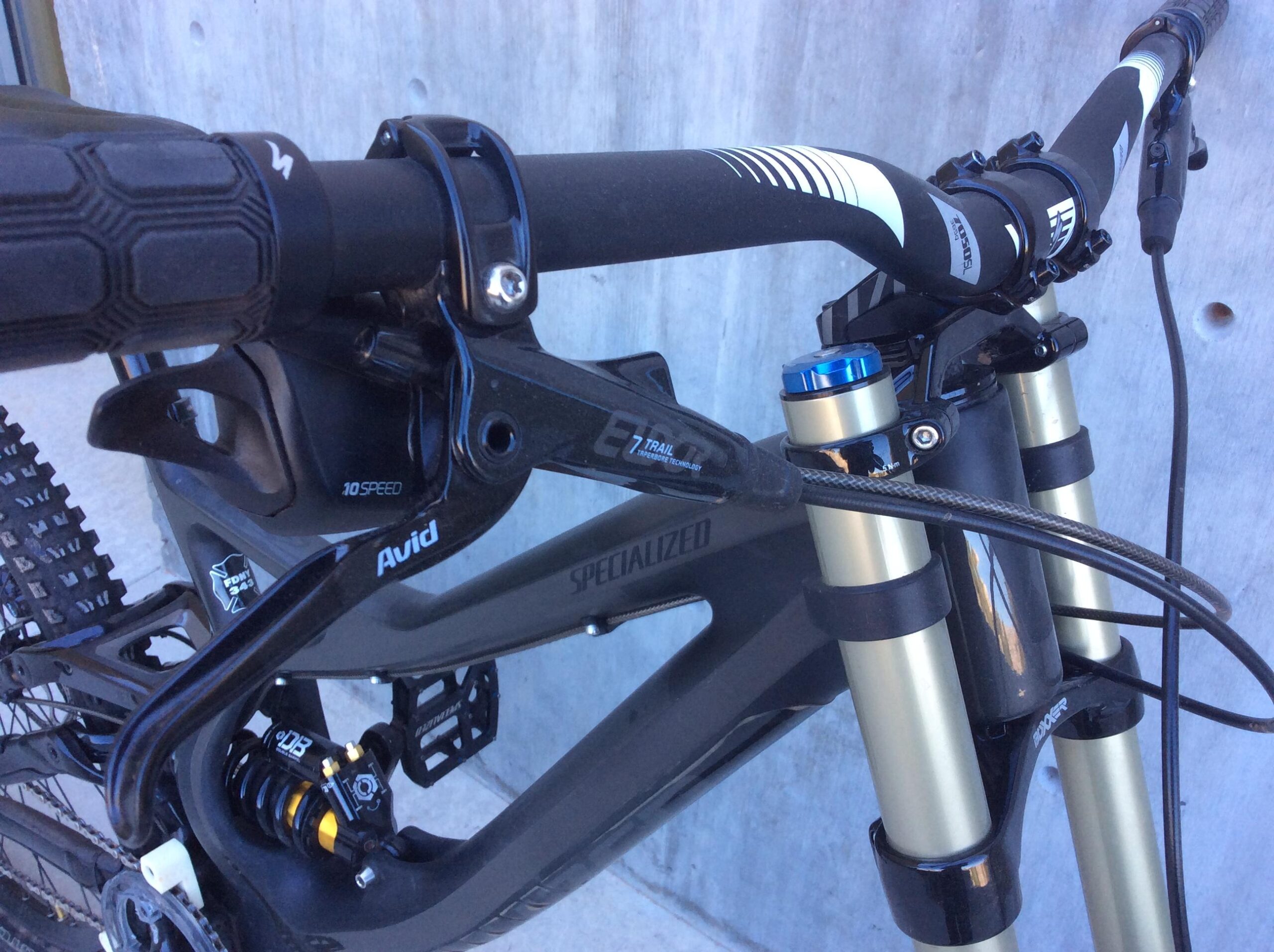 Specialized S-Works Demo 8 Carbon: Close-up view of a mountain bike's handlebars, featuring a black grip, gear shifter labeled "10 Speed," and a suspension fork with blue and gold components. The bike frame, branded "Specialized," is prominently displayed, alongside mechanical parts from Avid and a visible shock absorber. The background is a textured concrete surface.