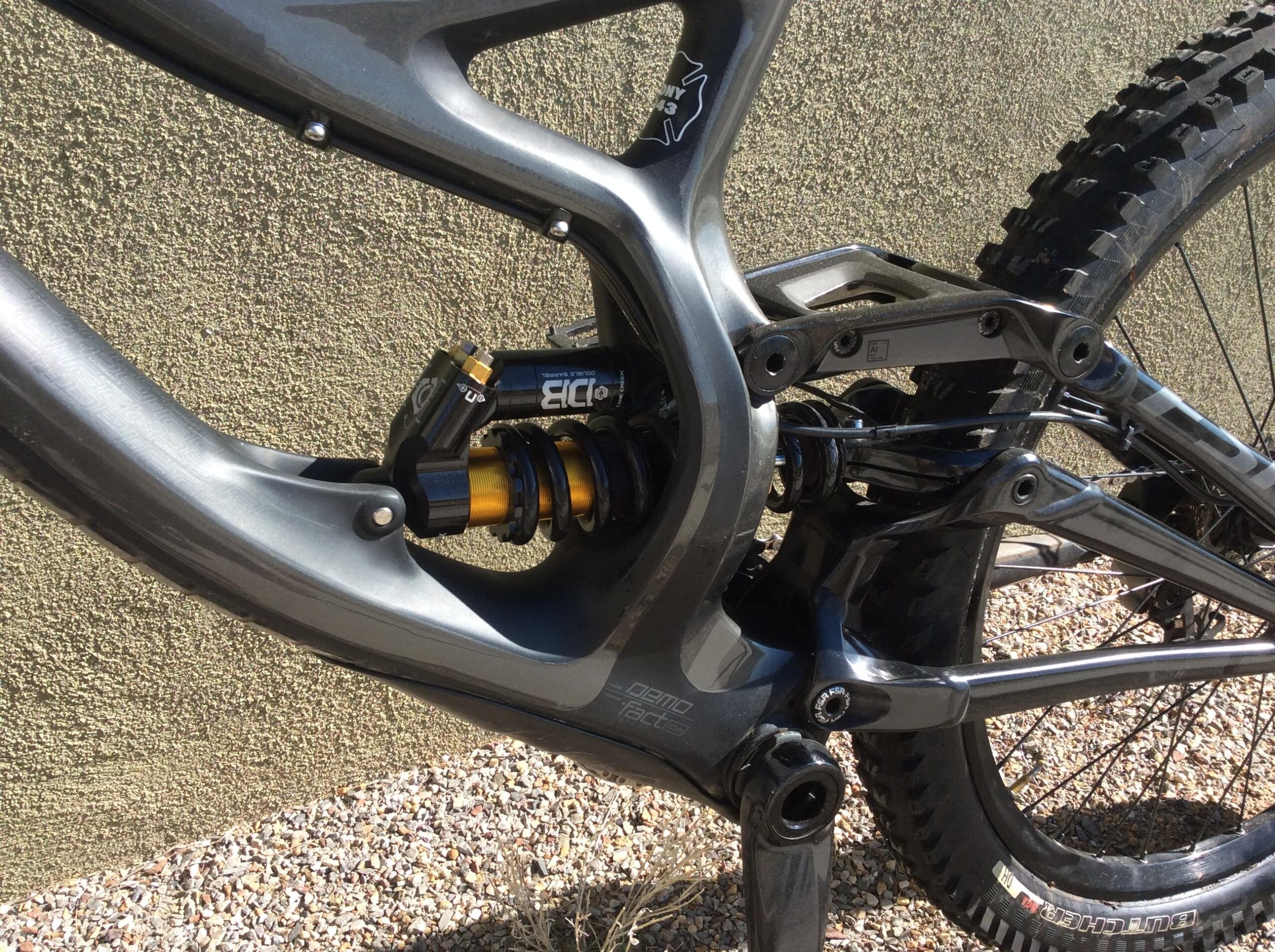 Specialized S-Works Demo 8: Close-up view of a mountain bike's rear suspension system, featuring a black and gold shock absorber integrated within the frame, set against a textured green wall and gravel surface.