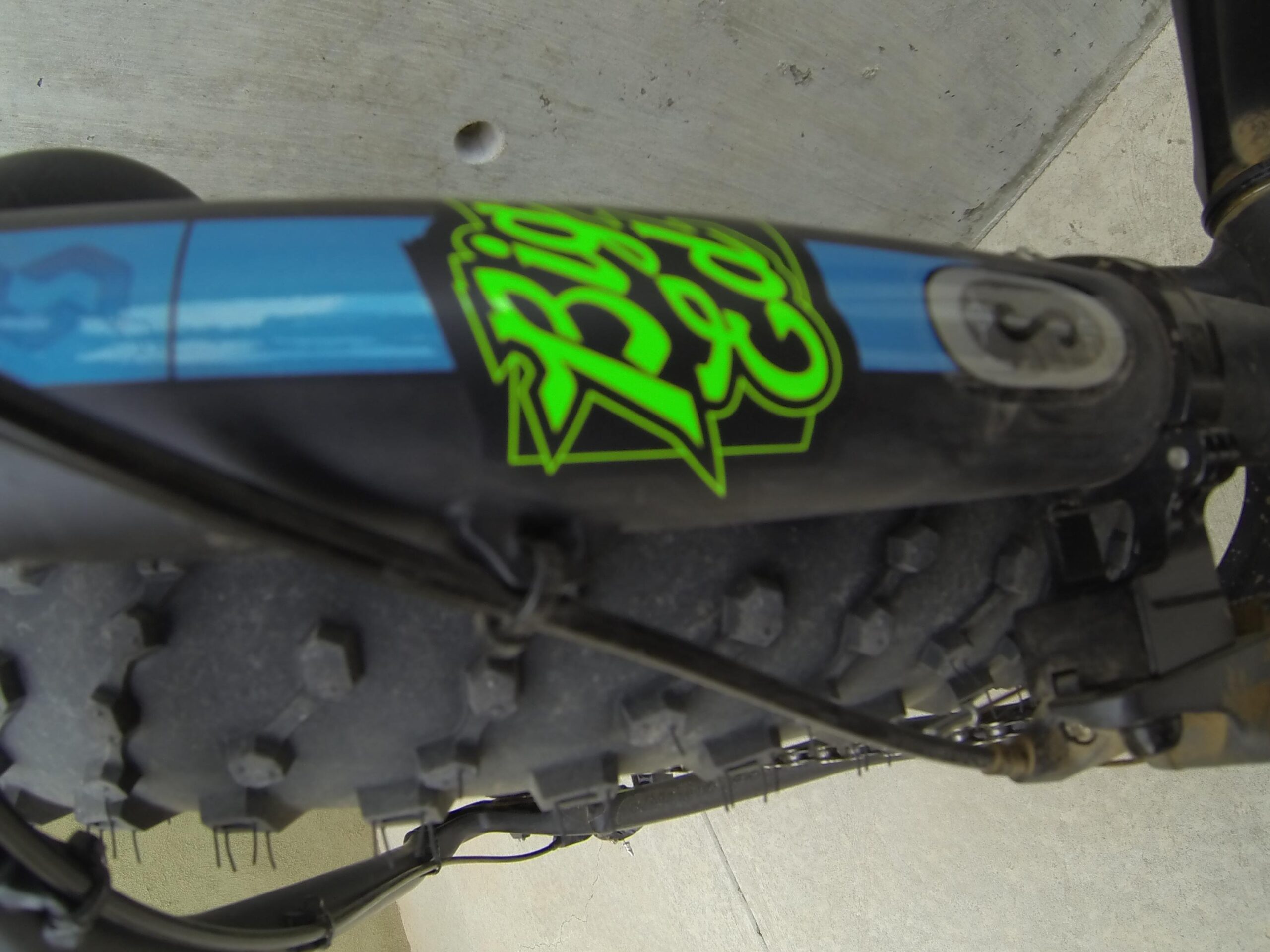 Scott Big Ed: Close-up view of a mountain bike frame featuring a blue and green design and a knobby tire. The bike frame is partially obscured, showing some cable housing and a gear mechanism. The surface underneath appears to be concrete.