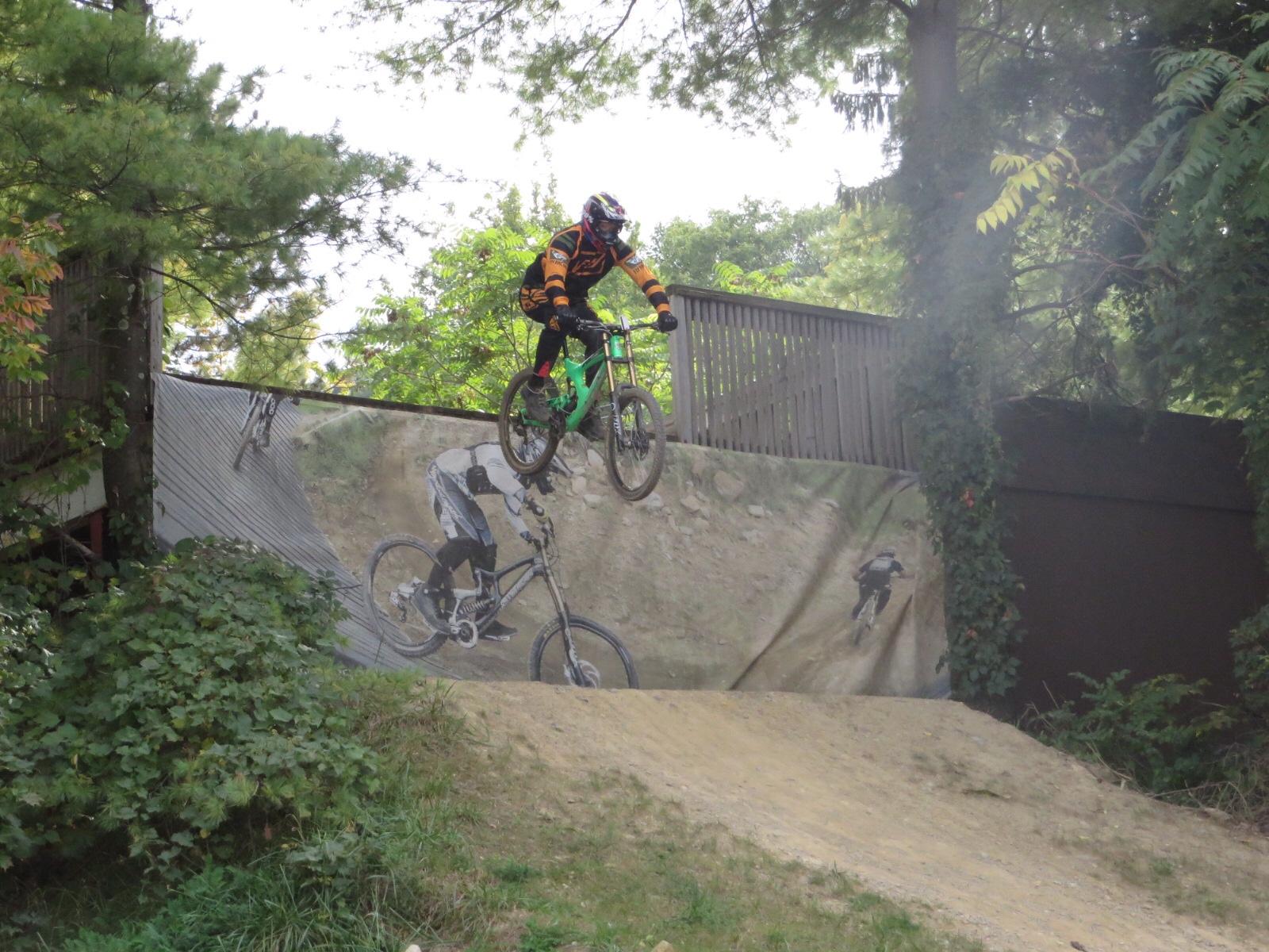 Giant Anthem: A mountain biker in an orange and black outfit performs a jump over a dirt ramp surrounded by trees, with a mural depicting another cyclist in the background.