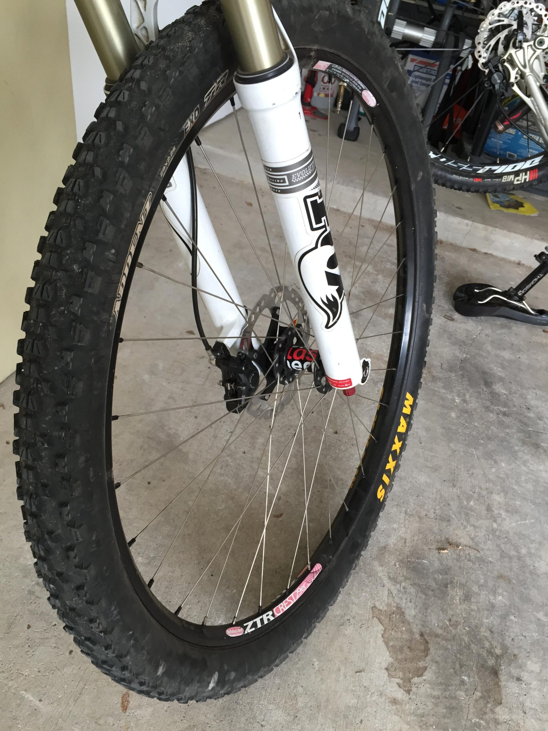 Trek Superfly 100 AL: Close-up view of a mountain bike front tire with a distinct tread pattern, mounted on a white suspension fork. The tire is branded Maxxis and features a ZTR Crest rim. The background shows a garage setting with tools and other bike parts visible.