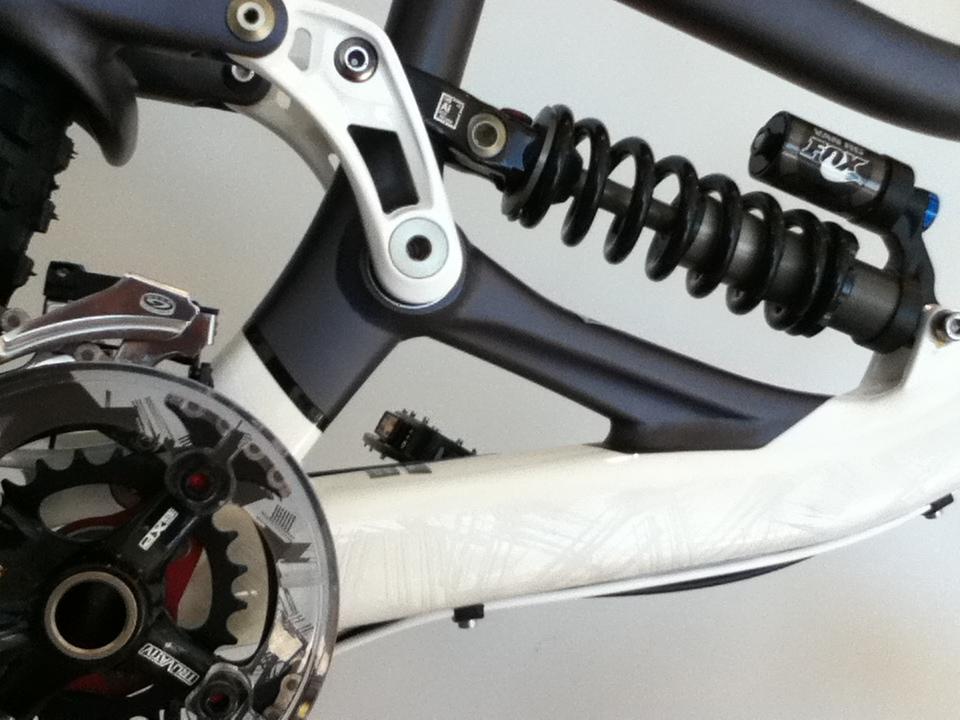Close-up view of a mountain bike's suspension system, showcasing the rear shock absorber, swingarm, and part of the drivetrain. The image focuses on the mechanical components, featuring a coiled spring shock, links, and the crankset with a detailed chainring. The frame has a predominantly black and white design with subtle graphics.