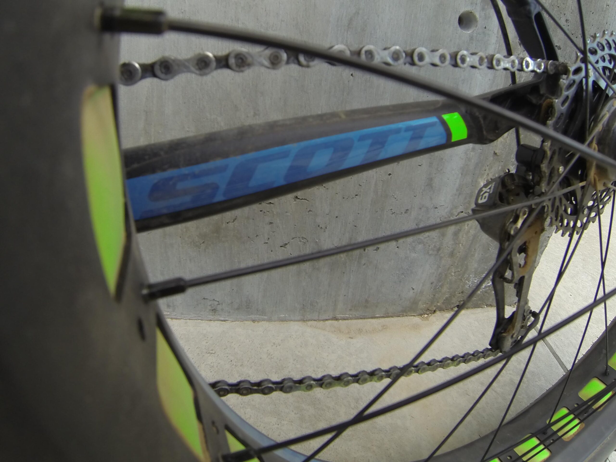 Scott Big Ed: Close-up view of a bicycle chain and rear derailleur, featuring a blue frame with the brand name "Scott" visible. The background shows a textured concrete surface, with part of the bike wheel and spokes in focus.