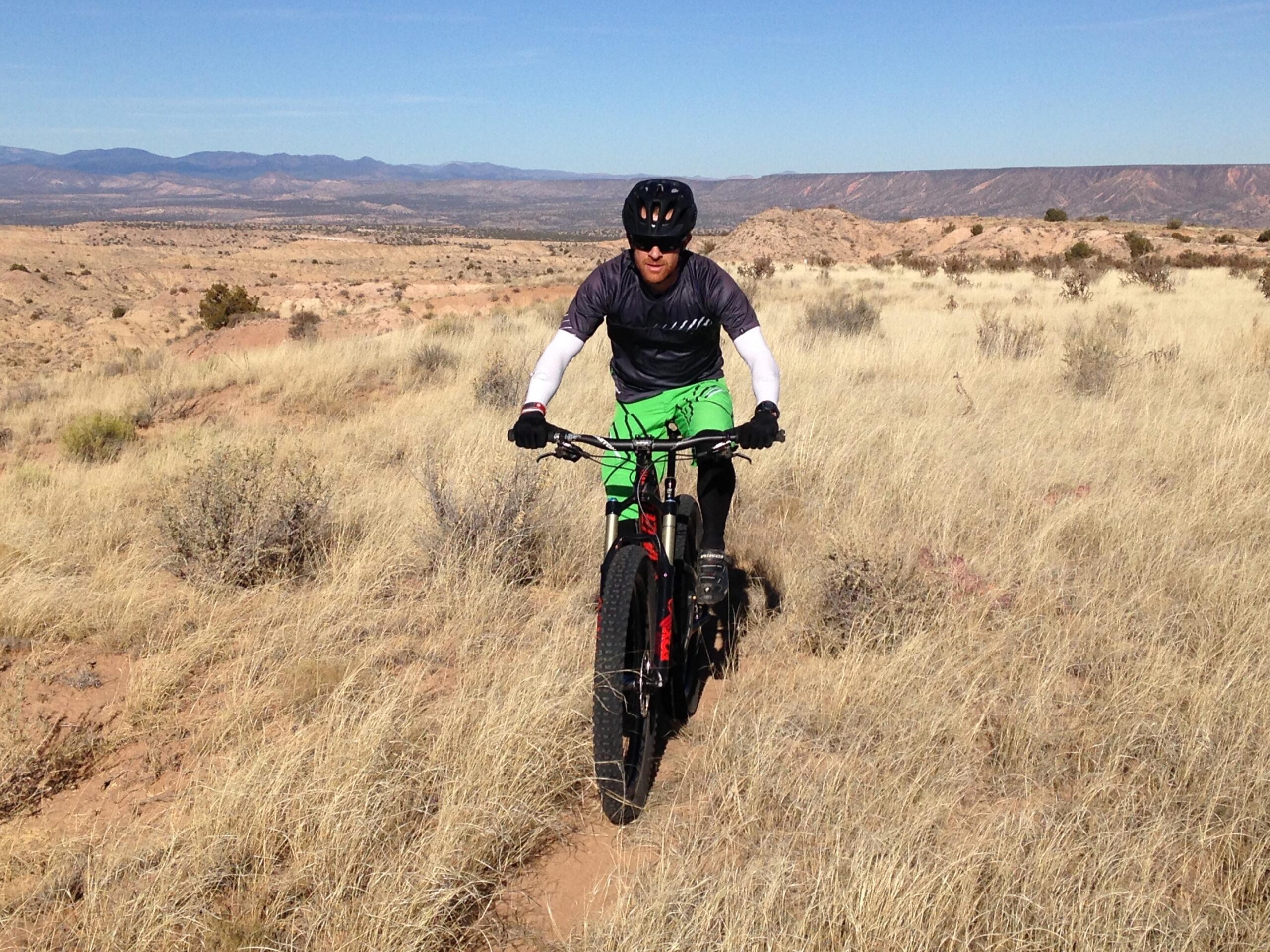 Specialized Stumpjumper FSR Pro: A person riding a mountain bike on a dirt trail surrounded by tall yellow grass, with a vast landscape of hills and mountains in the background under a clear blue sky. The cyclist is wearing a black helmet, a short-sleeve dark shirt, and bright green shorts.