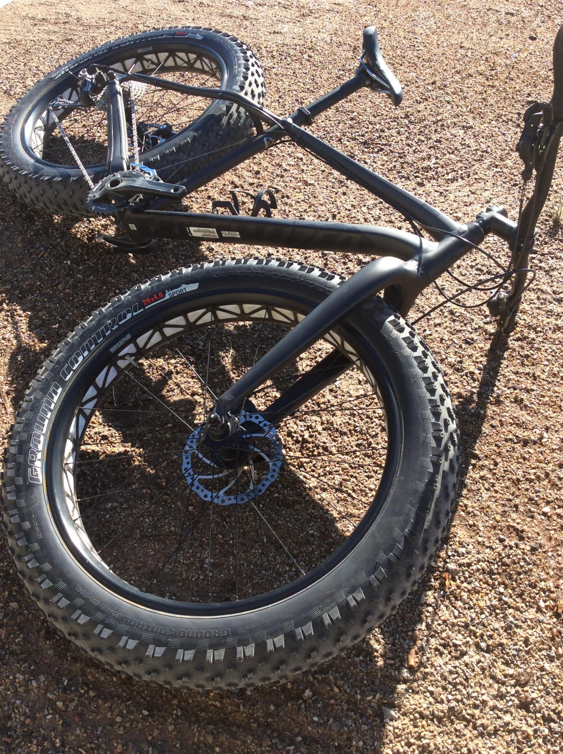 Specialized Fatboy: A close-up view of a black fat bike lying on a gravel surface, showcasing its large, textured tires and sturdy frame. The bike is positioned upside down, revealing its chainstay and wheel components, with sunlight highlighting the various parts.