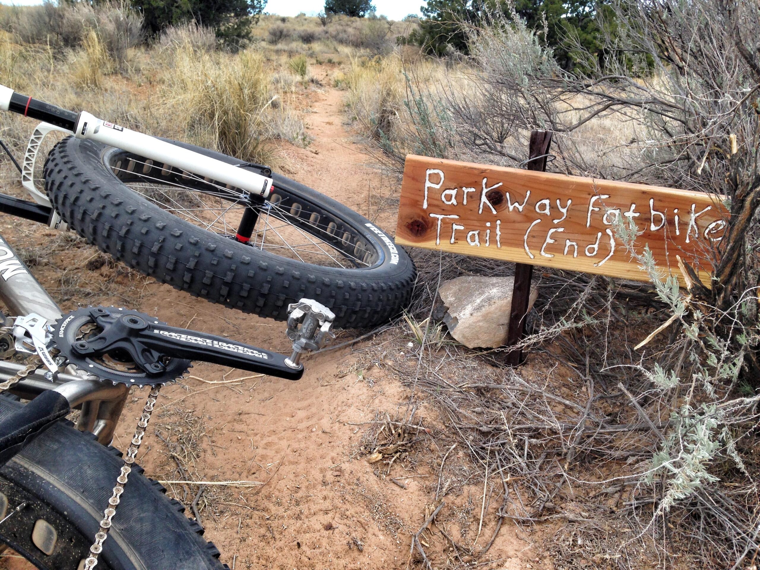 A close-up view of a fatbike with a large tire resting on sandy terrain next to a wooden trail sign that reads "Parkway Fatbike Trail (End)." The background features sparse vegetation and a dirt path leading into the distance. Parkway Fatbike trail mountain bike trail.