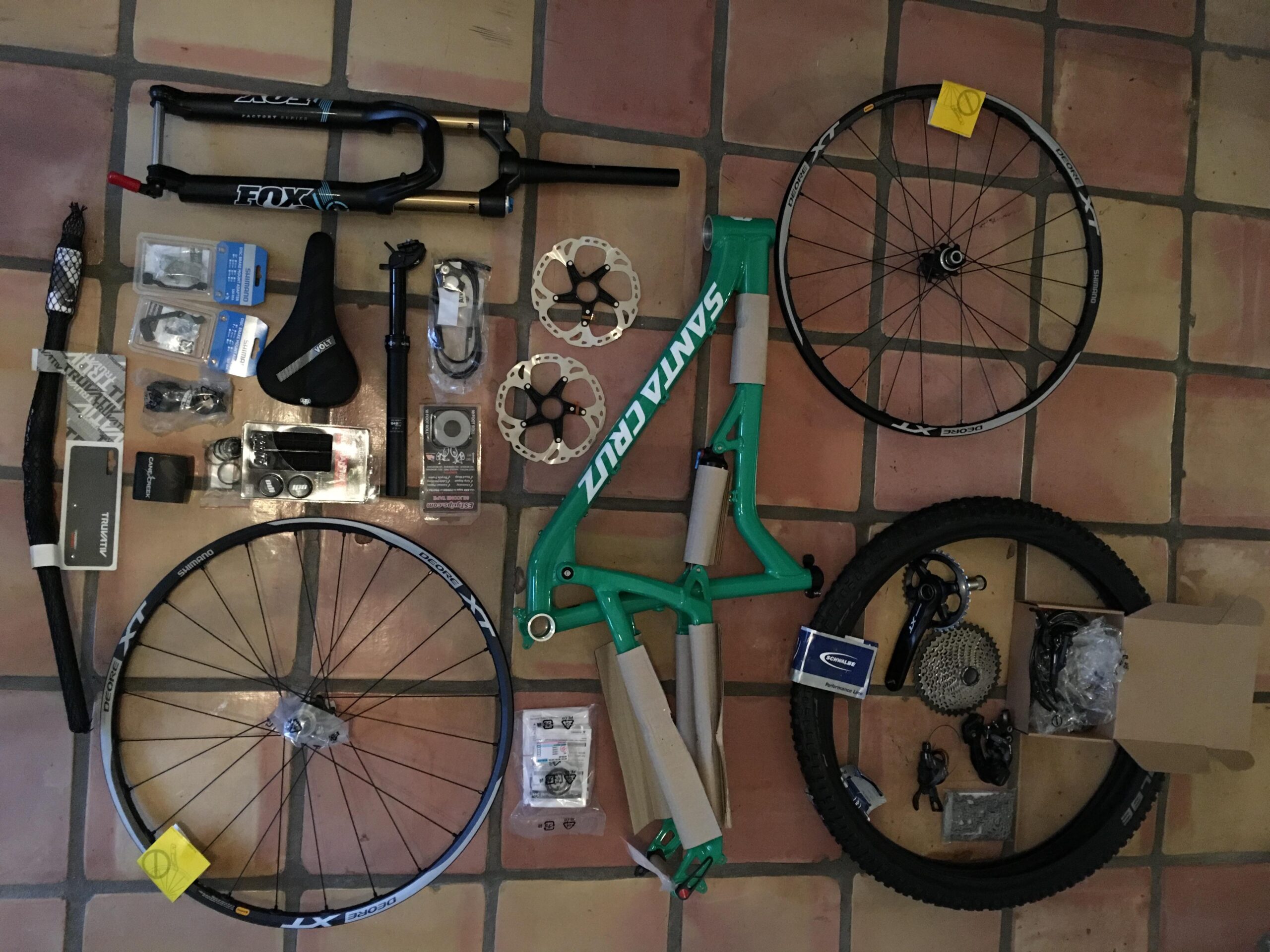 Santa Cruz Bantam: Image of various mountain bike components arranged on a tiled surface. Items include a green bike frame, two wheels, a fork, a bicycle saddle, brake rotors, tools, and a selection of other bike parts and accessories.