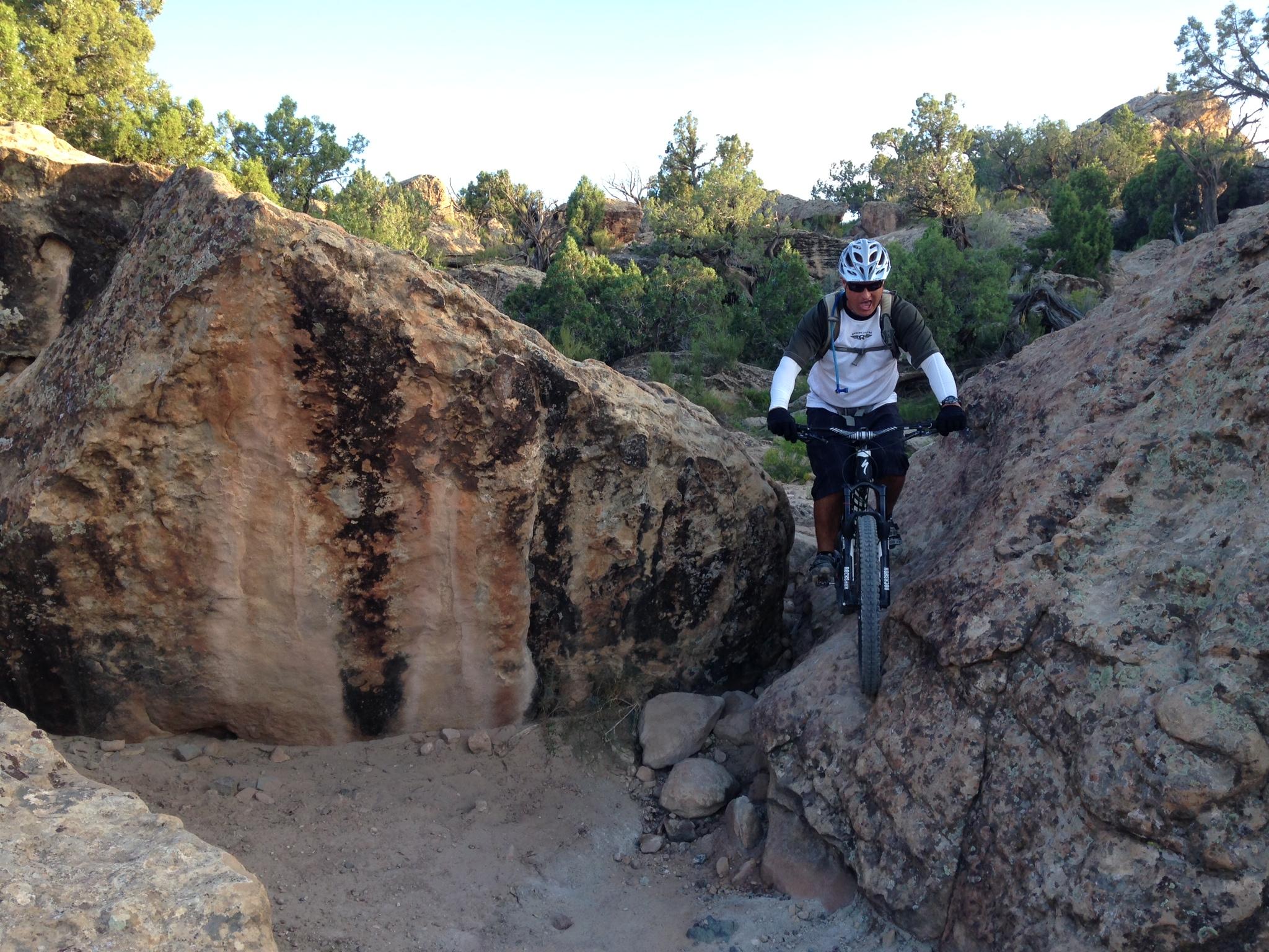 Specialized Enduro S-Works 650b: A mountain biker navigating a narrow passage between large rocks in a natural outdoor setting, with greenery and trees in the background.