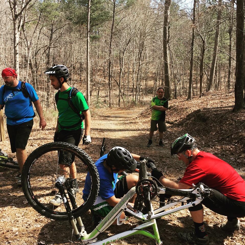 A group of five cyclists, dressed in colorful jerseys, is gathered on a dirt trail in a wooded area. Two individuals are focused on repairing a bicycle, while the others observe. The environment features bare trees and fallen leaves, indicating an early spring setting. Uwharrie NF: Wood Run, Supertree And Keyauwee mountain bike trail.