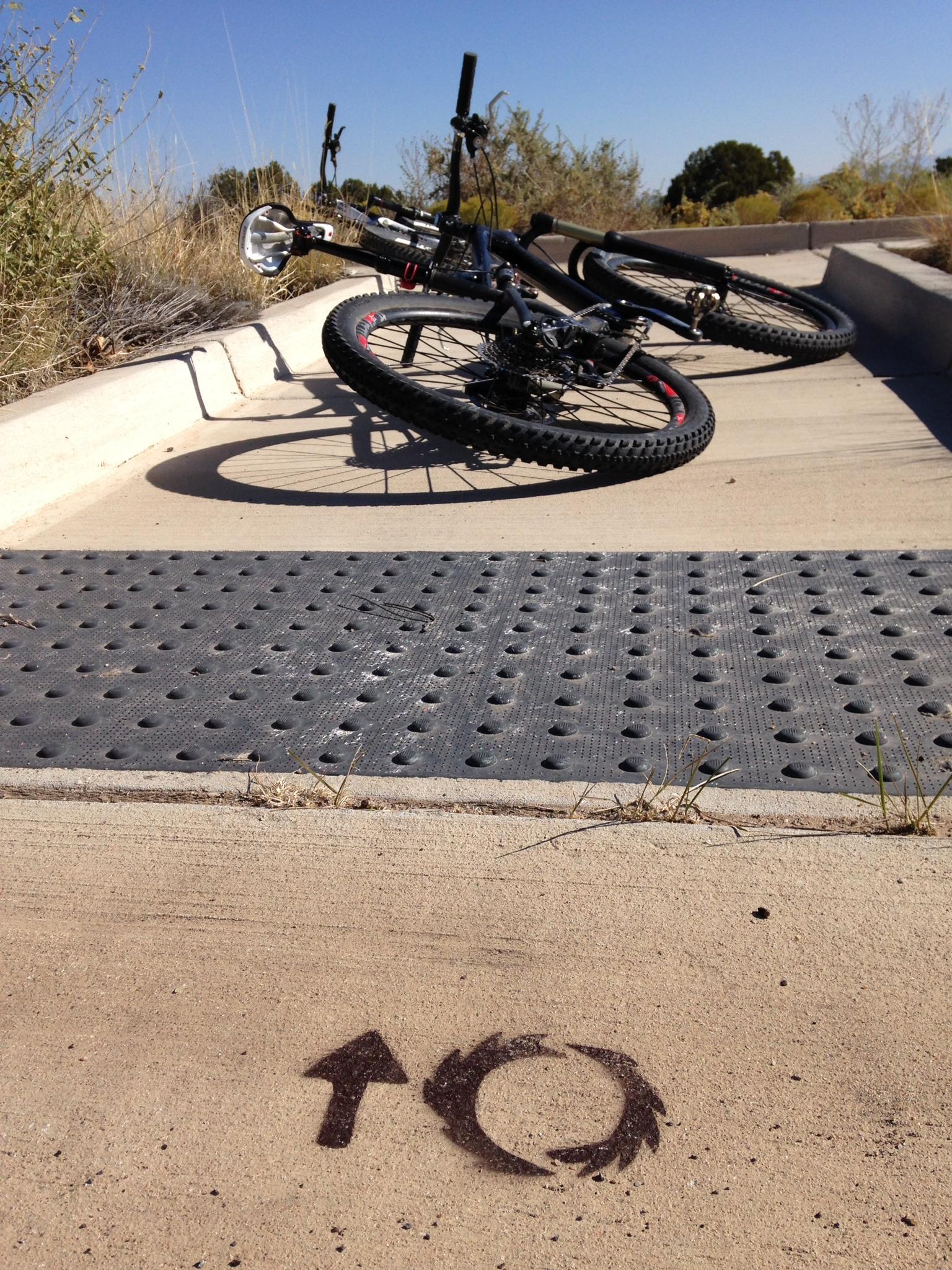 A close-up view of two mountain bikes resting on a concrete path, with the bike tires leaning against the side. In the foreground, a concrete surface features a directional arrow pointing upward and a circular symbol indicating a turn or loop. The background includes dry vegetation and bright blue sky, suggesting an outdoor location. Parkway Fatbike trail mountain bike trail.