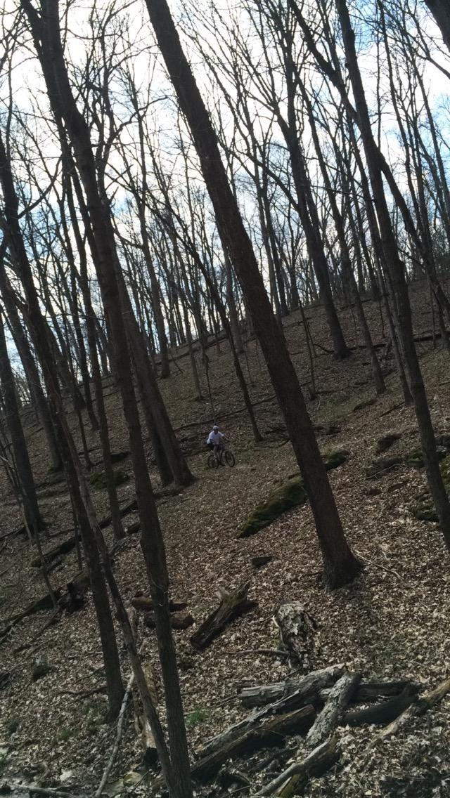 A mountain biker riding through a wooded area with bare trees and scattered leaves on the ground, under a cloudy sky. Lost Valley mountain bike trail.