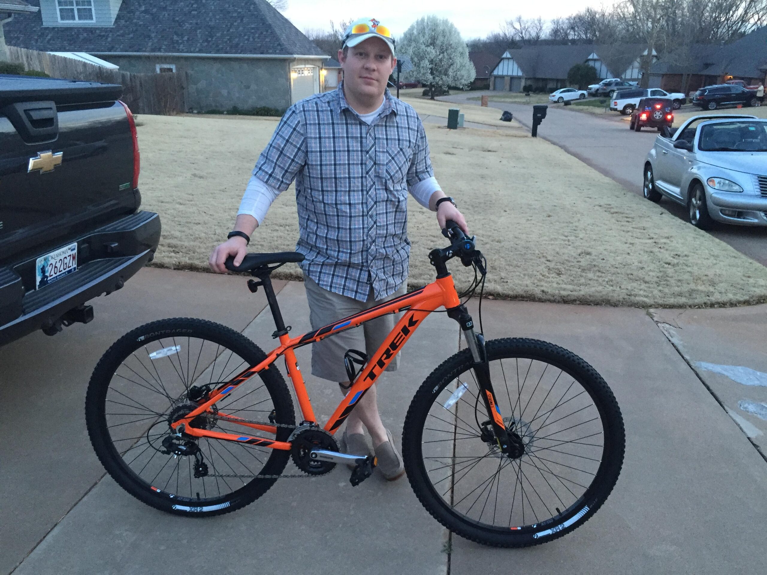 Trek Marlin 6: A person stands beside a bright orange Trek mountain bike on a suburban driveway, wearing a plaid shirt and light-colored shorts. The bike features thick tires and a sturdy frame, with a Chevrolet truck visible in the background. The setting is an early evening with grass and houses lining the street.