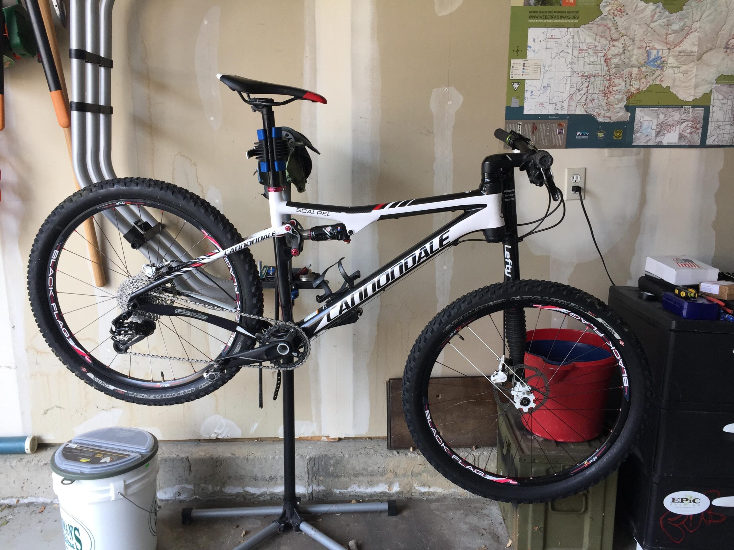 Cannondale Scalpel: A mountain bike suspended on a repair stand in a garage, with tools and a map visible in the background. The bike features a white and black frame, marked with "Scalpel" and "Cannondale," and has large tires with "Black Flag" branding. The garage has a concrete floor and storage containers nearby.