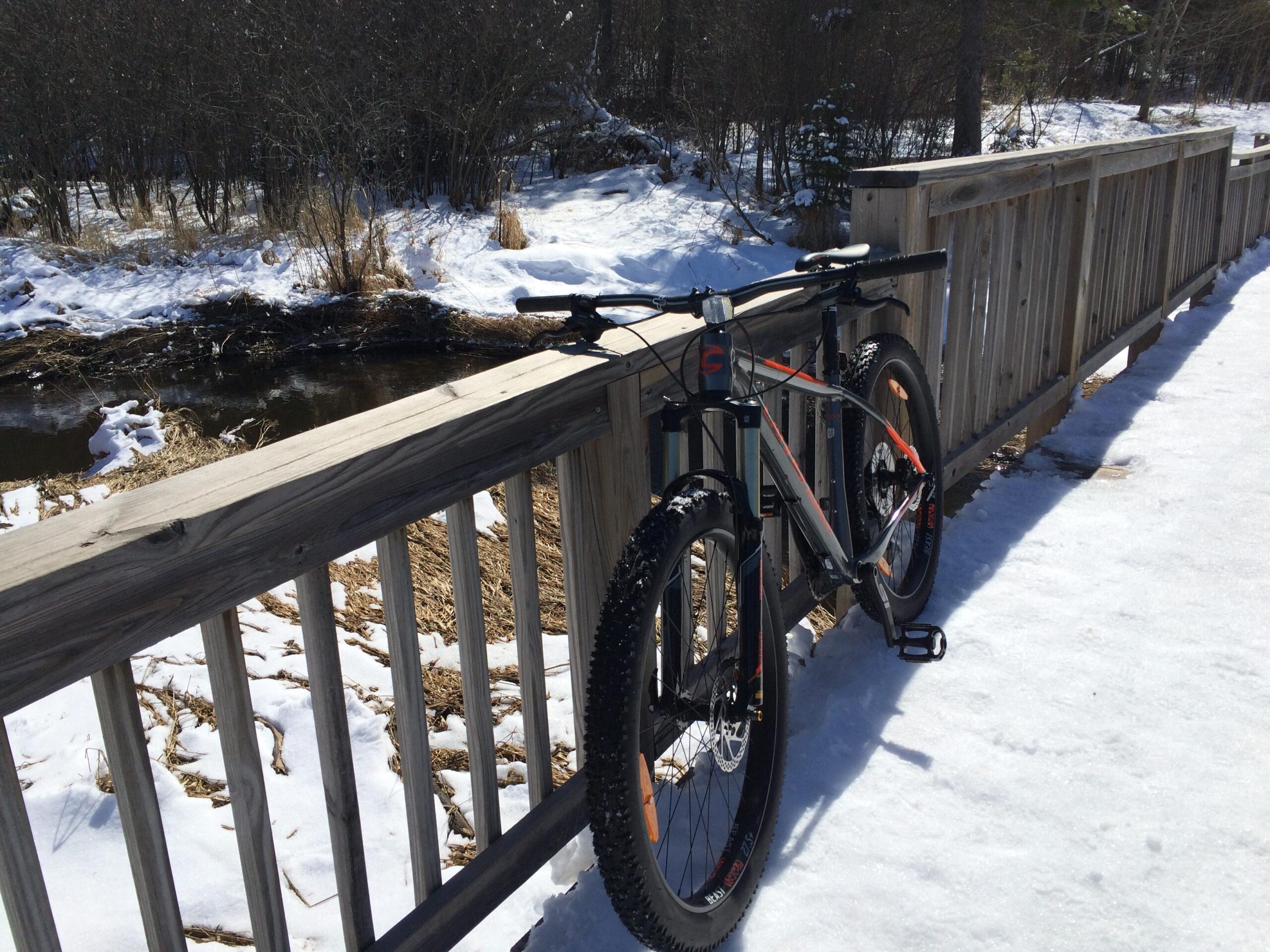 A mountain bike leaning against a wooden railing, overlooking a snow-covered landscape with patches of grass and a small stream visible in the background. The scene is bright and sunny, showcasing a winter setting. Hartley Park mountain bike trail.