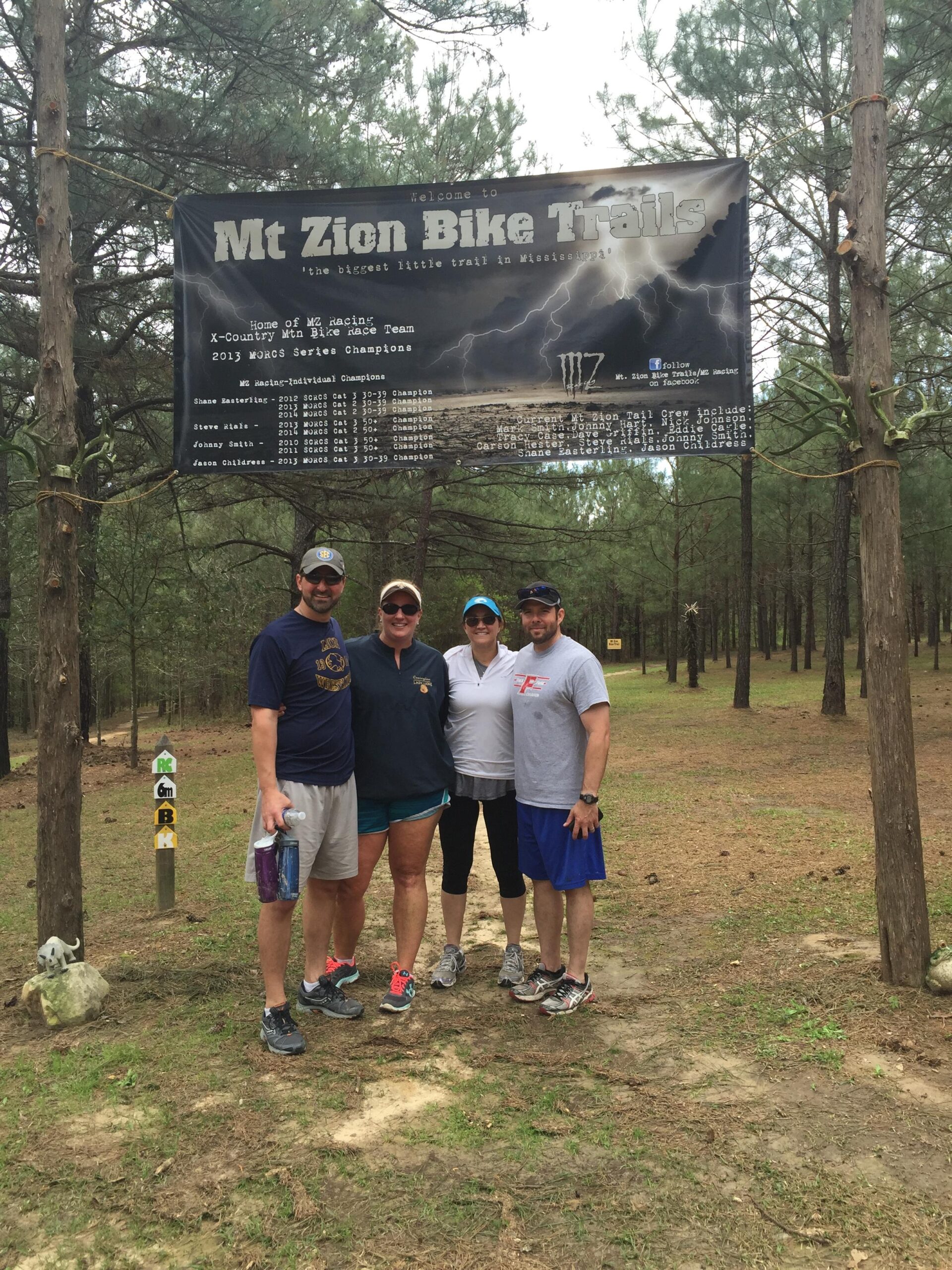 A group of four people stands in front of a large sign that welcomes visitors to Mt Zion Bike Trails, which promotes itself as "the biggest little trail in Mississippi." The scene is set in a wooded area with tall trees and a dirt path visible in the background. The group is smiling, wearing casual outdoor clothing. Beside them, there are colorful trail markers indicating different bike routes. Mt. Zion Bike Trails mountain bike trail.