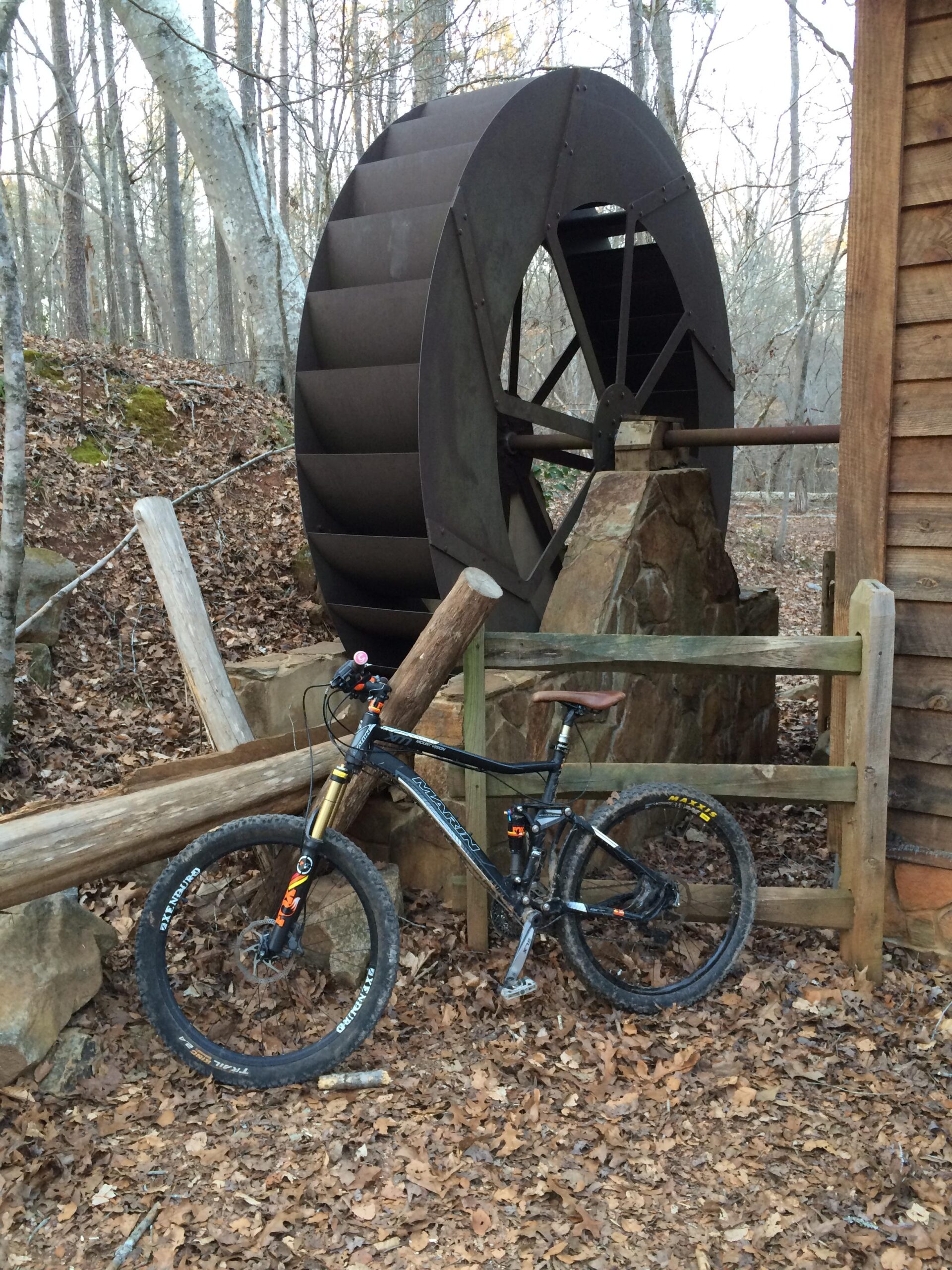 Alt text: A mountain bike rests on a bed of fallen leaves next to a large, metal water wheel and a wooden structure in a forested area. Anne Springs Close Greenway mountain bike trail.
