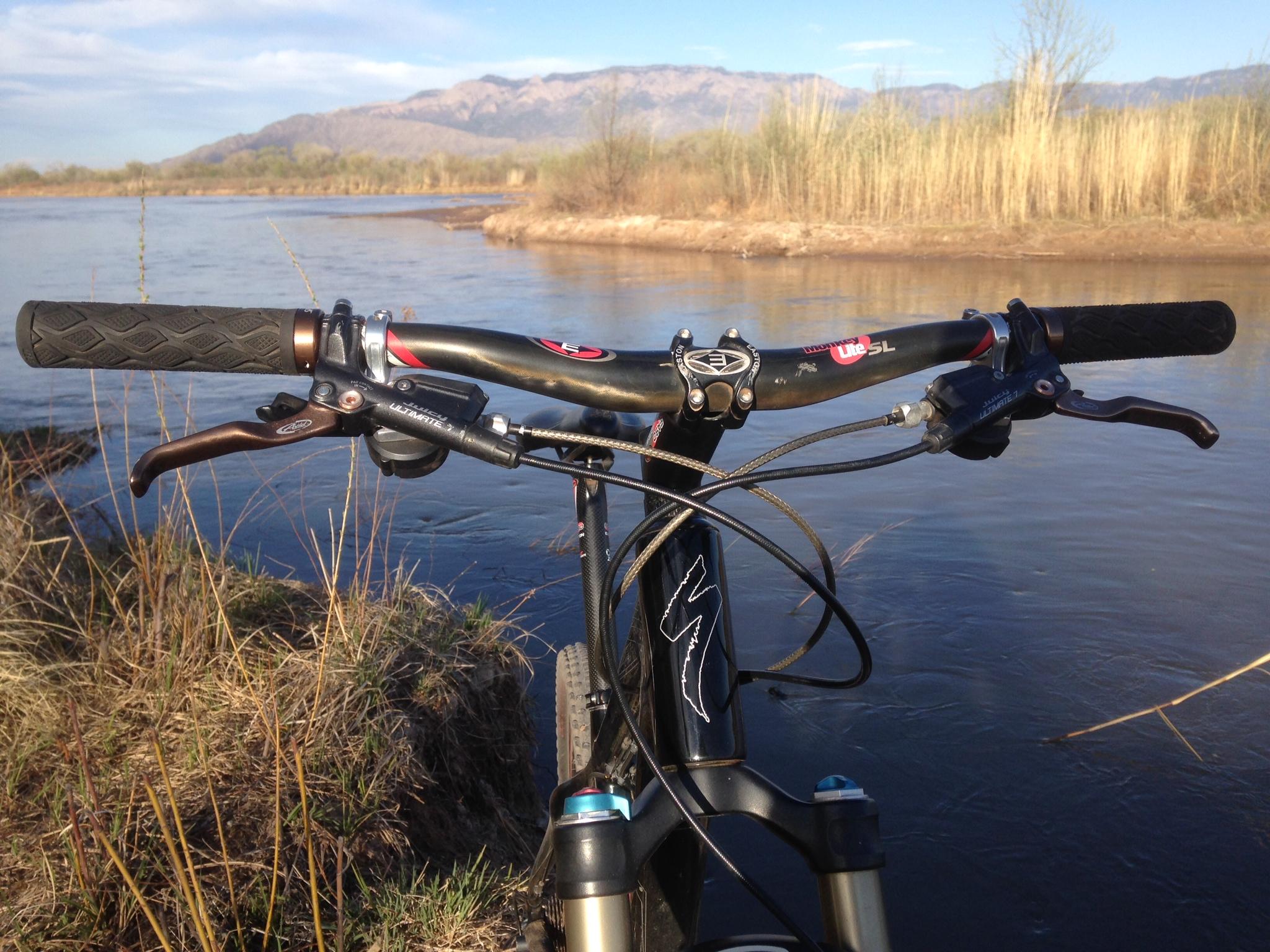 A close-up view of a mountain bike's handlebars positioned near a riverbank, with a scenic backdrop of mountains and trees. The sun is shining, and the water reflects the blue sky. Albuquerque Bosque mountain bike trail.
